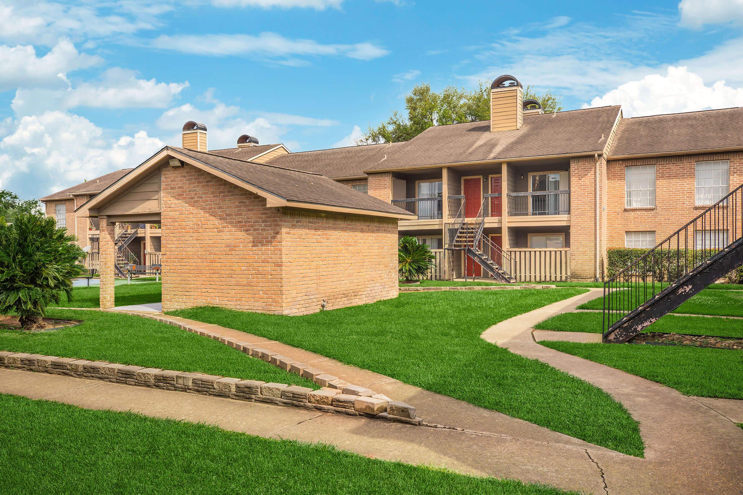A landscaped residential apartment complex featuring brick buildings, green grass, and stone pathways. The scene includes two buildings with balconies, a small separate structure, and a decorative walkway under a blue sky with clouds.