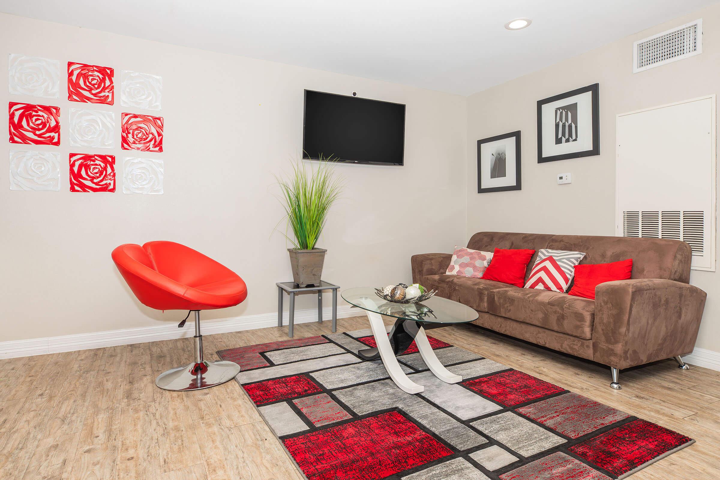 A modern living room featuring a brown sofa with red and white pillows, a stylish red chair, a glass coffee table on a patterned rug, and decorative wall art, including red roses. A flat-screen TV is mounted on the wall, and a potted plant adds a touch of greenery.