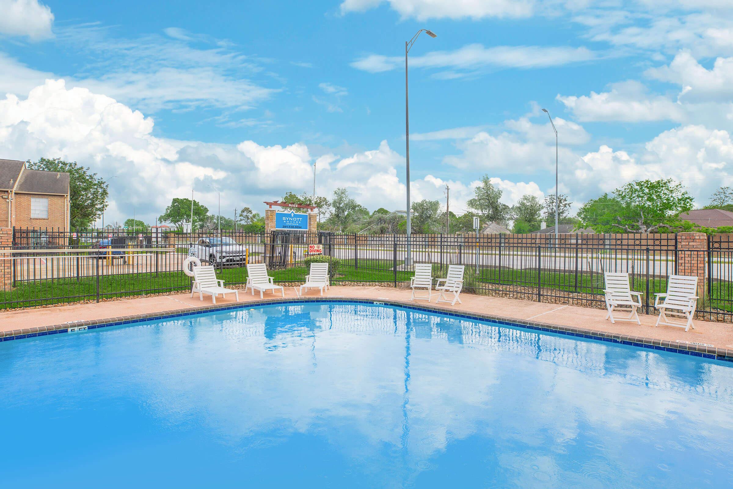 A clear swimming pool surrounded by white lounge chairs and a fence. Lush green grass and trees are visible in the background, with a blue sky and fluffy white clouds above. There is a sign in the background indicating the pool area, and a road visible beyond the fence.