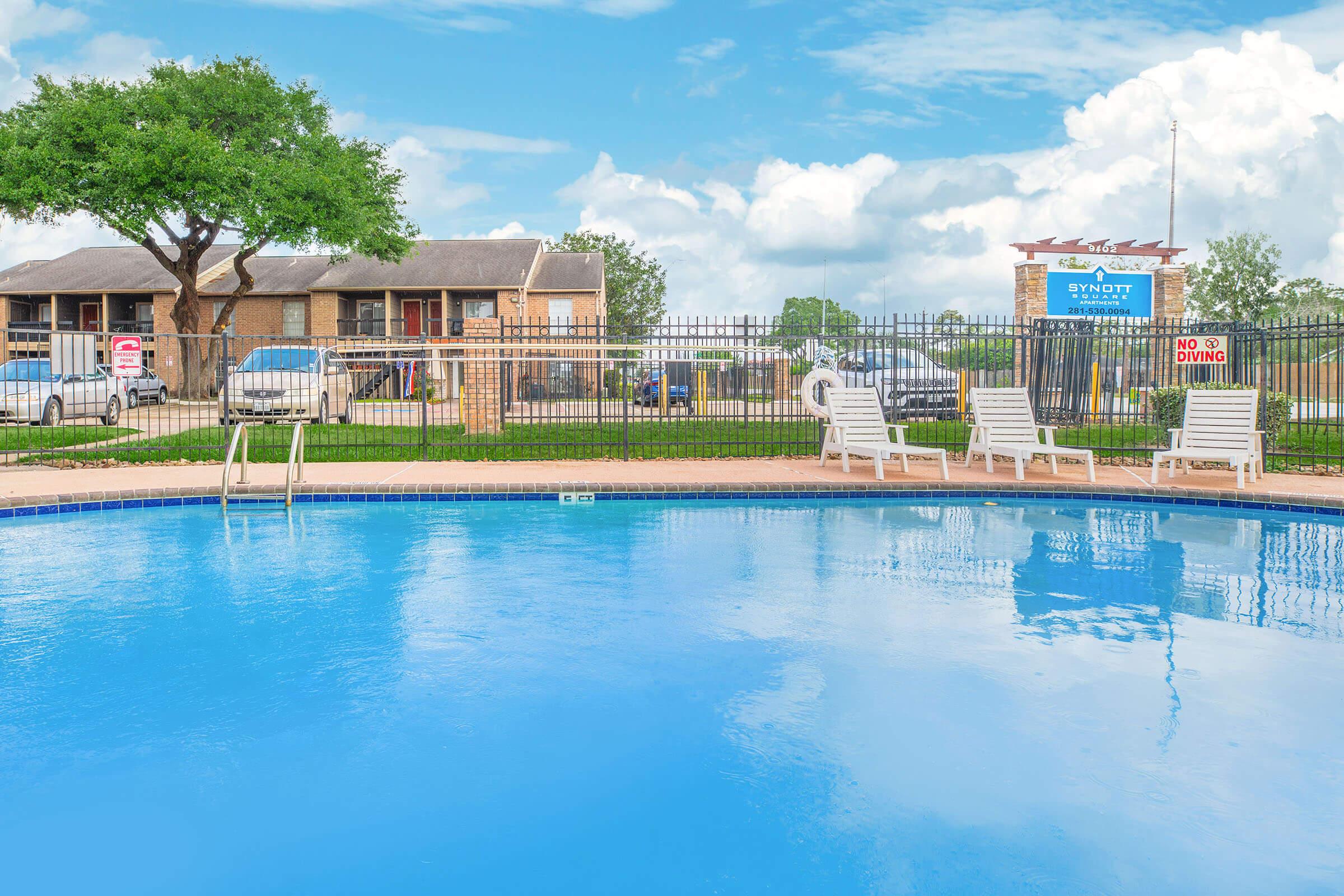 A clear blue swimming pool surrounded by a fenced area, with several empty lounge chairs positioned nearby. In the background, a two-story building and sign for "Syndott" can be seen, along with parked cars and a tree under a partly cloudy sky. No people are present.