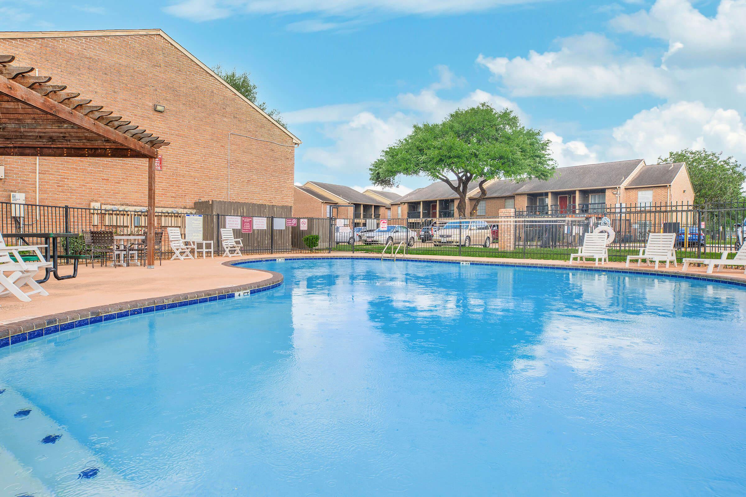 A bright and inviting outdoor swimming pool surrounded by lounge chairs and a fenced patio area. In the background, there are brick buildings and lush green trees under a clear blue sky. The scene conveys a relaxing and recreational atmosphere.