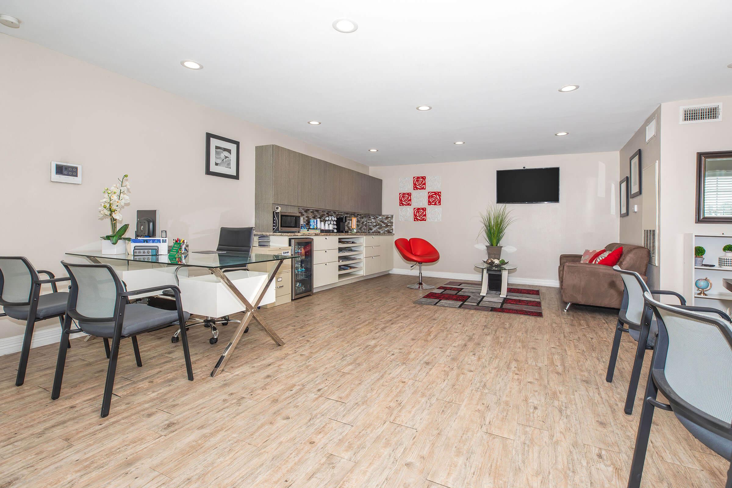 Modern office space featuring a wooden floor, a central coffee station, and a glass conference table with black chairs. A cozy seating area includes a red accent chair and a brown couch. The walls are adorned with decorative art, and a flat-screen TV is mounted for viewing. Natural light fills the room, creating an inviting atmosphere.