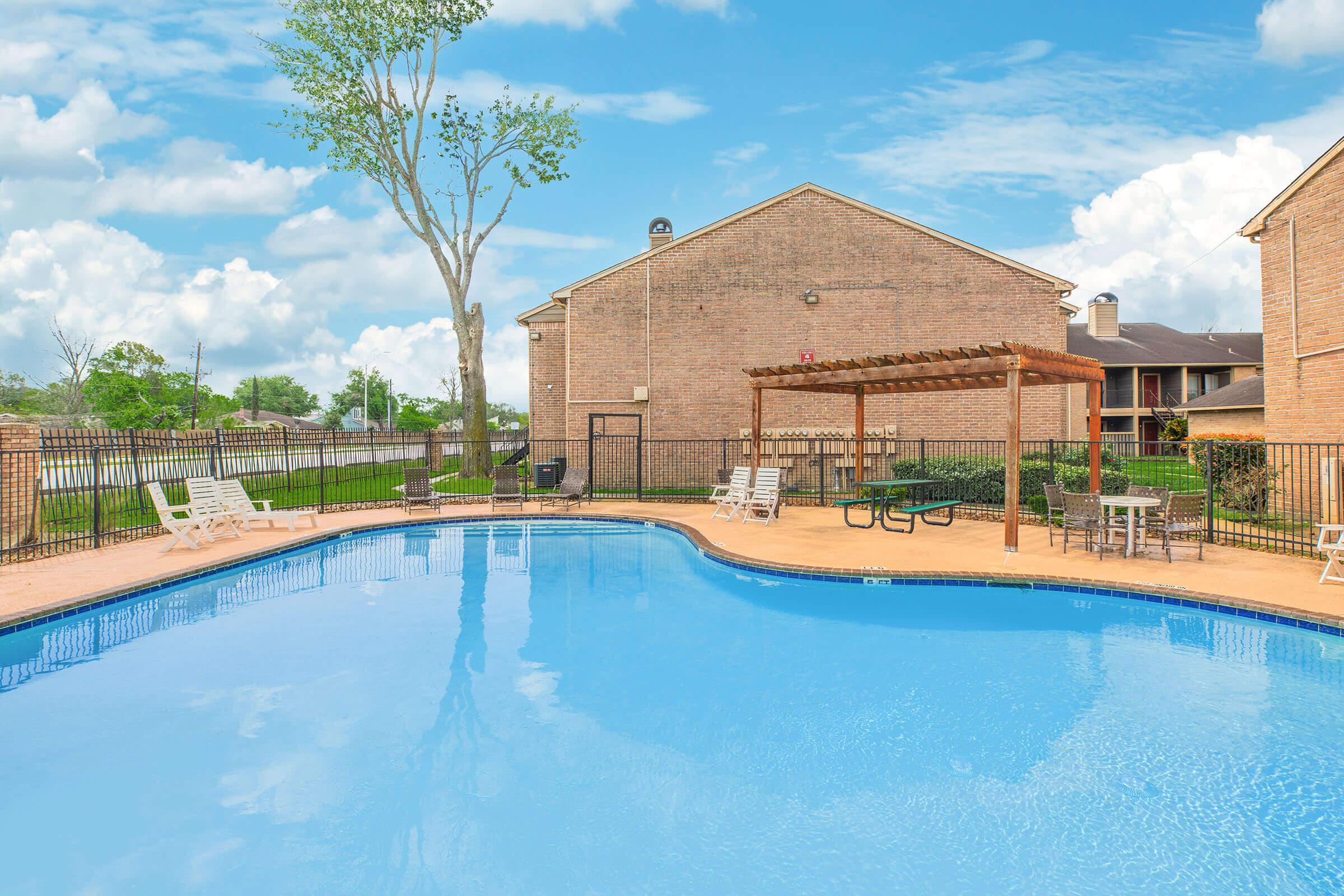 A clear blue swimming pool surrounded by lounge chairs, a shaded picnic area with tables, and a pergola. In the background, there are two brick buildings and a tree, under a bright sky with fluffy clouds. The scene conveys a peaceful and inviting atmosphere for relaxation and leisure.