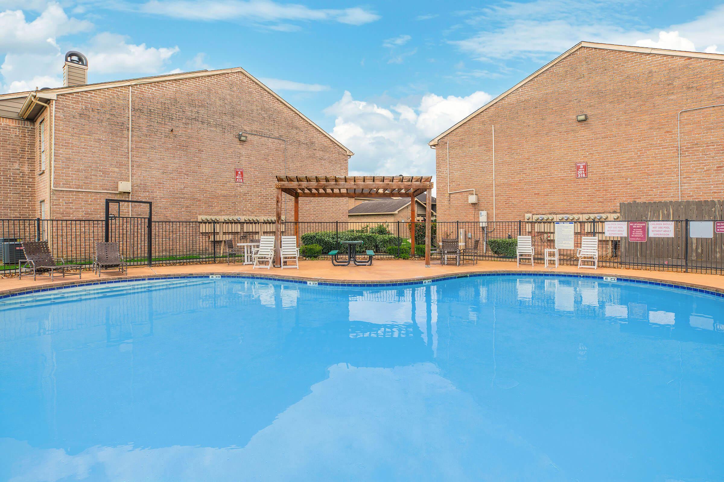 A clear blue swimming pool surrounded by a wooden fence, lounge chairs, and a pergola. In the background, two brick residential buildings are visible, against a bright blue sky with scattered clouds. The area appears well-maintained, inviting relaxation and leisure.
