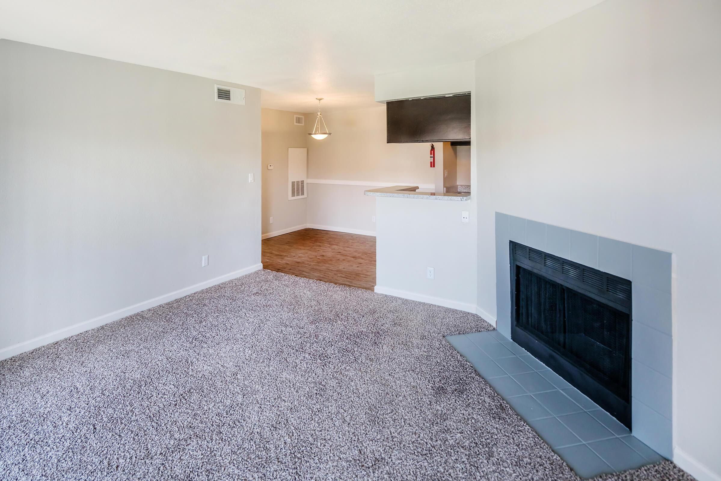 A spacious living area with light gray walls and plush, beige carpeting, featuring a fireplace with a gray surround. To the back, there is an open kitchen with a breakfast bar, natural lighting from a nearby window, and a dining area with a pendant light above.