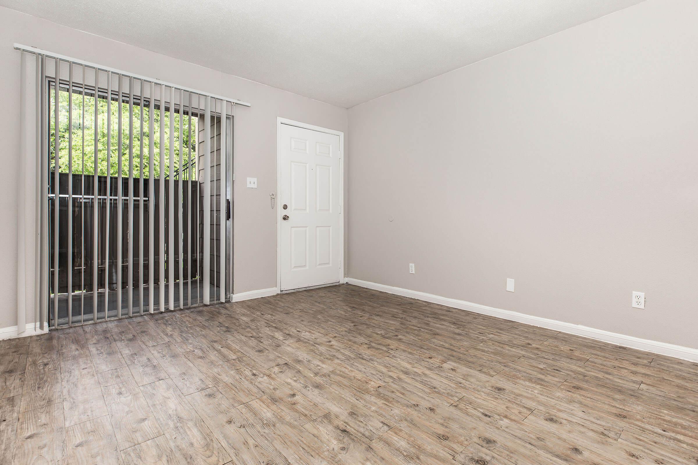 Empty room with light gray walls and a wooden floor. A door on the left leads to a small balcony, which features vertical blinds. The room is well-lit and spacious, with a single white door in the center and no furniture.