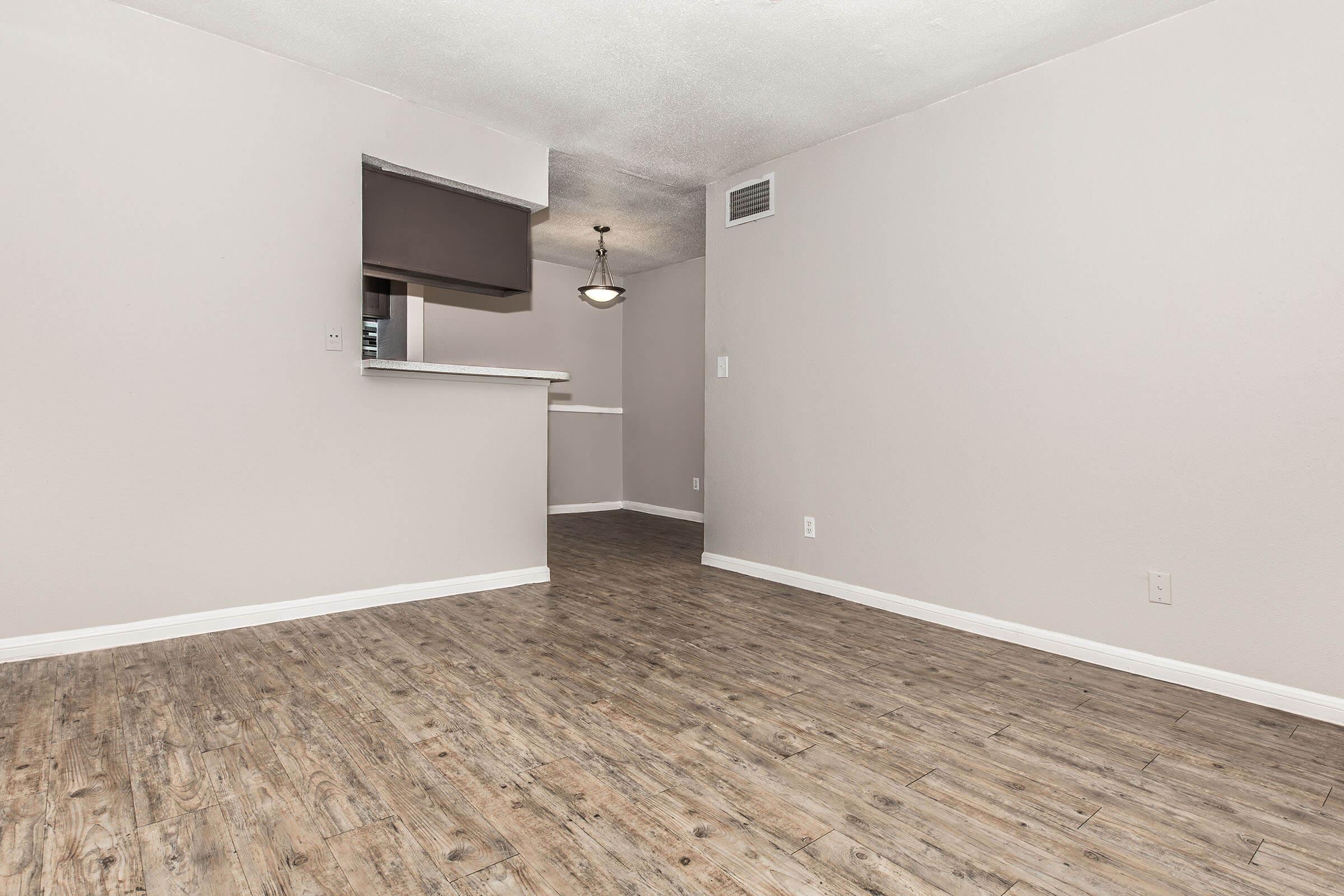 A spacious, light-colored room with wooden flooring, featuring a small kitchen area visible in the background. The walls are painted in a soft shade, and there is a light fixture hanging from the ceiling. The area is empty, creating an open and inviting atmosphere.