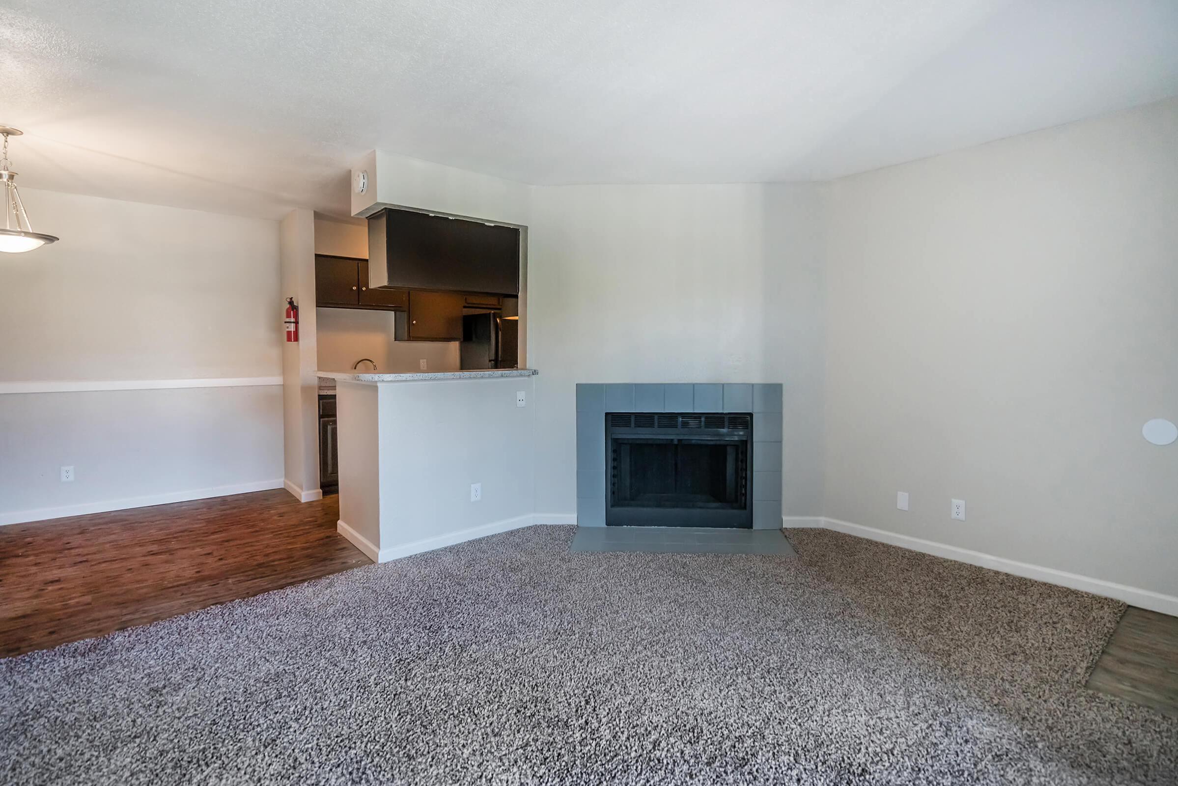 A spacious living area featuring plush carpet and a gray fireplace. To the left, a glimpse of a kitchen area with dark cabinetry. Walls are painted a light color, creating a bright and airy atmosphere. A pendant light hangs above, adding warmth to the space.
