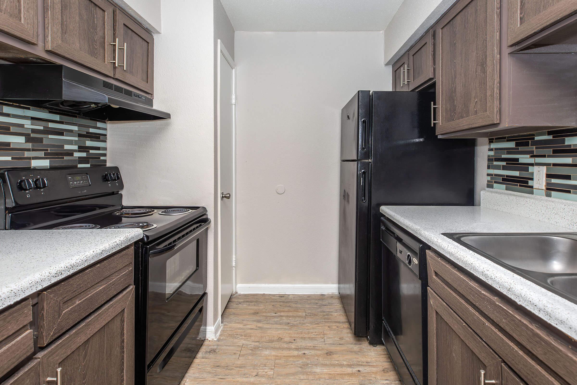 Modern kitchen featuring dark wooden cabinetry, a black refrigerator, an electric stove, and dual sinks. The walls are accented with a stylish backsplash of teal and brown tiles. The space has a clean and organized appearance with a wooden floor, providing a contemporary look.
