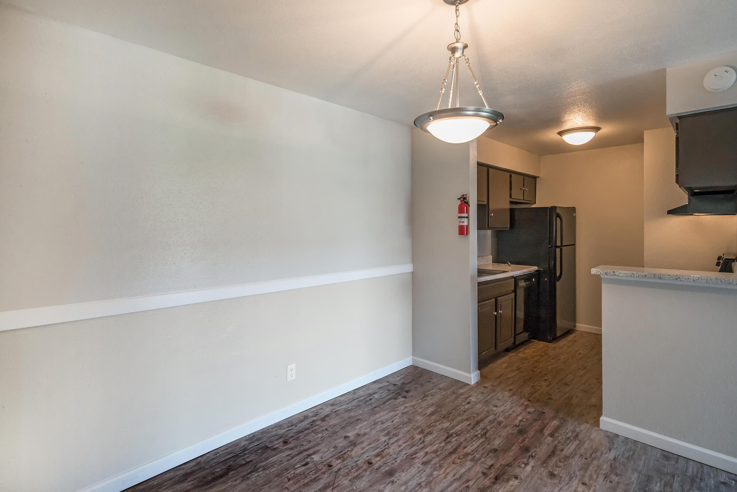 A well-lit kitchen area featuring grey cabinets, a refrigerator, and a countertop. The flooring is laminate wood. A pendant light hangs from the ceiling, with a fire extinguisher mounted on the wall. The walls are painted in neutral tones, enhancing the spacious feel of the room.