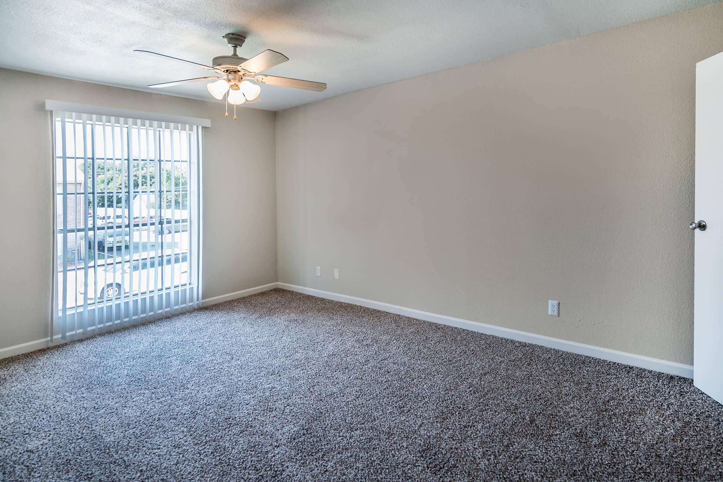 A spacious, empty room with light beige walls and plush carpeting. A ceiling fan is mounted in the center, and there are large windows covered with white vertical blinds, allowing natural light to brighten the space. A door on the right leads to another area.