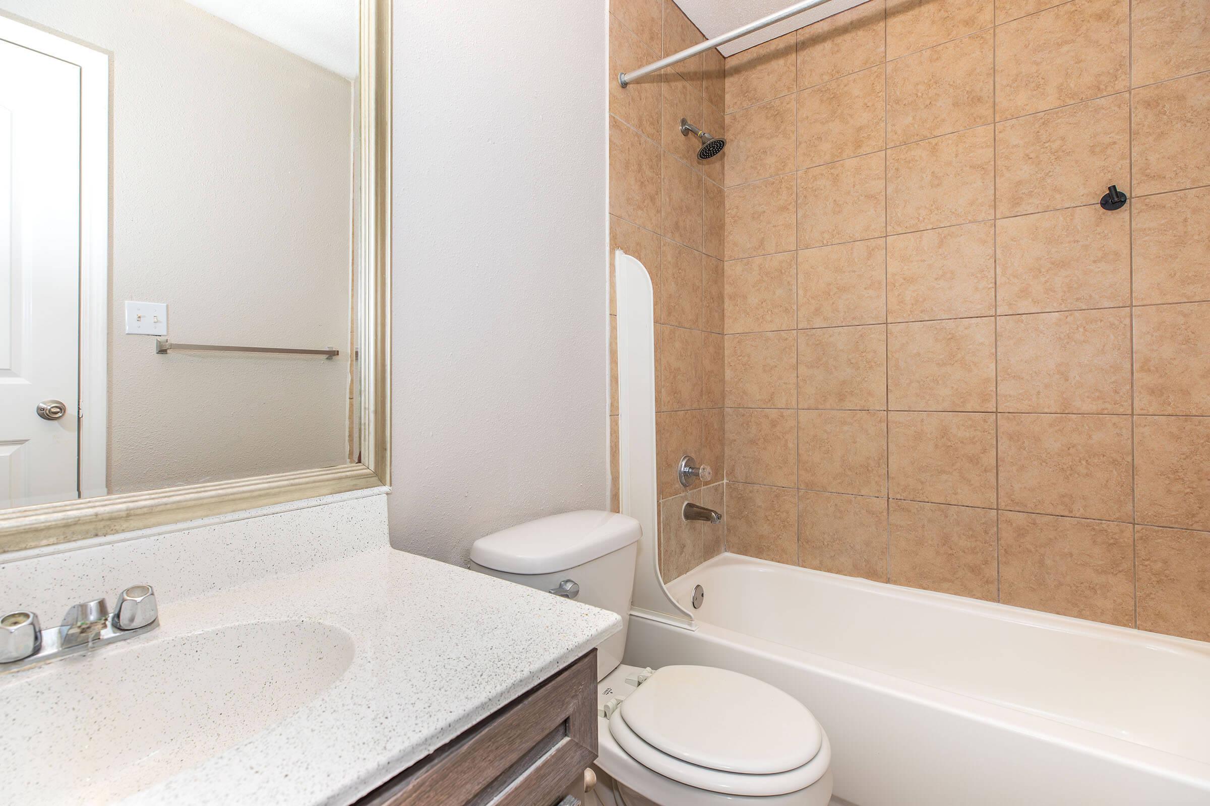 A clean bathroom featuring a bathtub, shower, and a toilet. The walls are covered in beige tiles, and there is a mirror above a countertop with a sink. A towel rack is mounted on the wall near the door. The overall atmosphere is bright and well-maintained.
