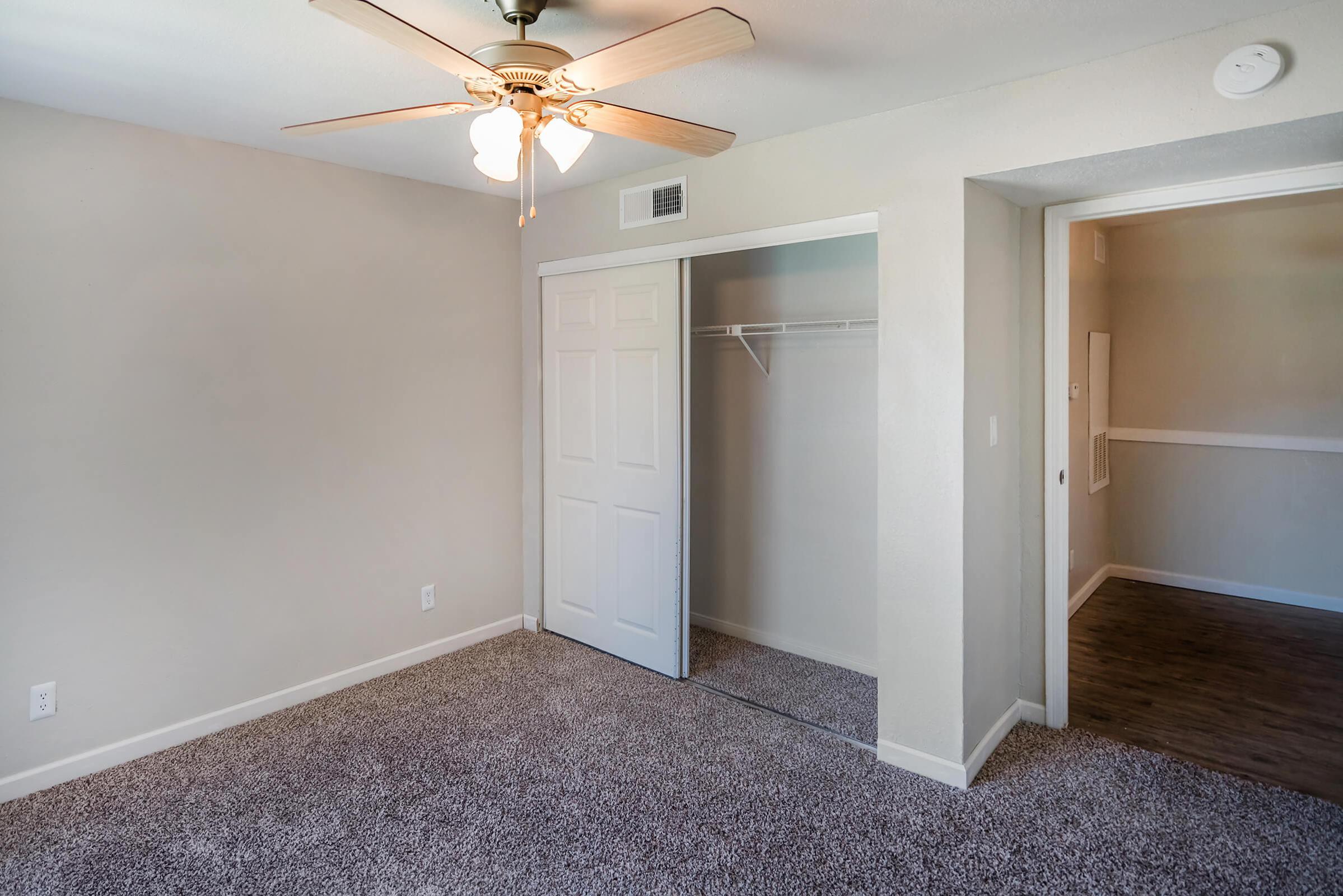 A well-lit room with beige walls and a ceiling fan. The floor is covered in light-colored carpet. On one side, there's a closet with mirrored sliding doors, and on the other side, an open hallway leading to another space. The overall ambiance is clean and inviting.