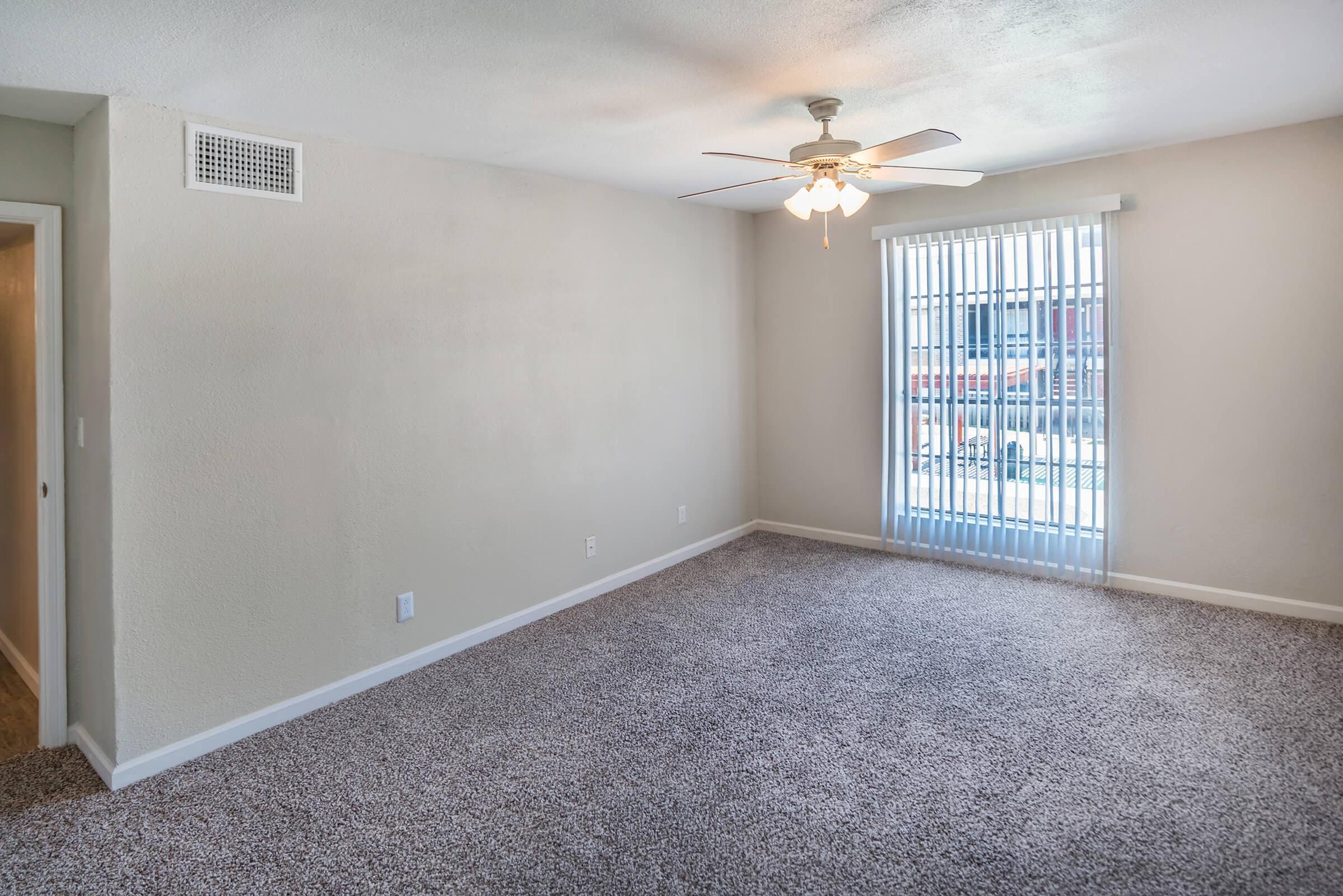A well-lit, empty living room with light beige walls and large vertical blinds covering a bright window. The ceiling features a white ceiling fan, and the floor is covered with soft, light-colored carpet. A doorway leads to another room on the left.