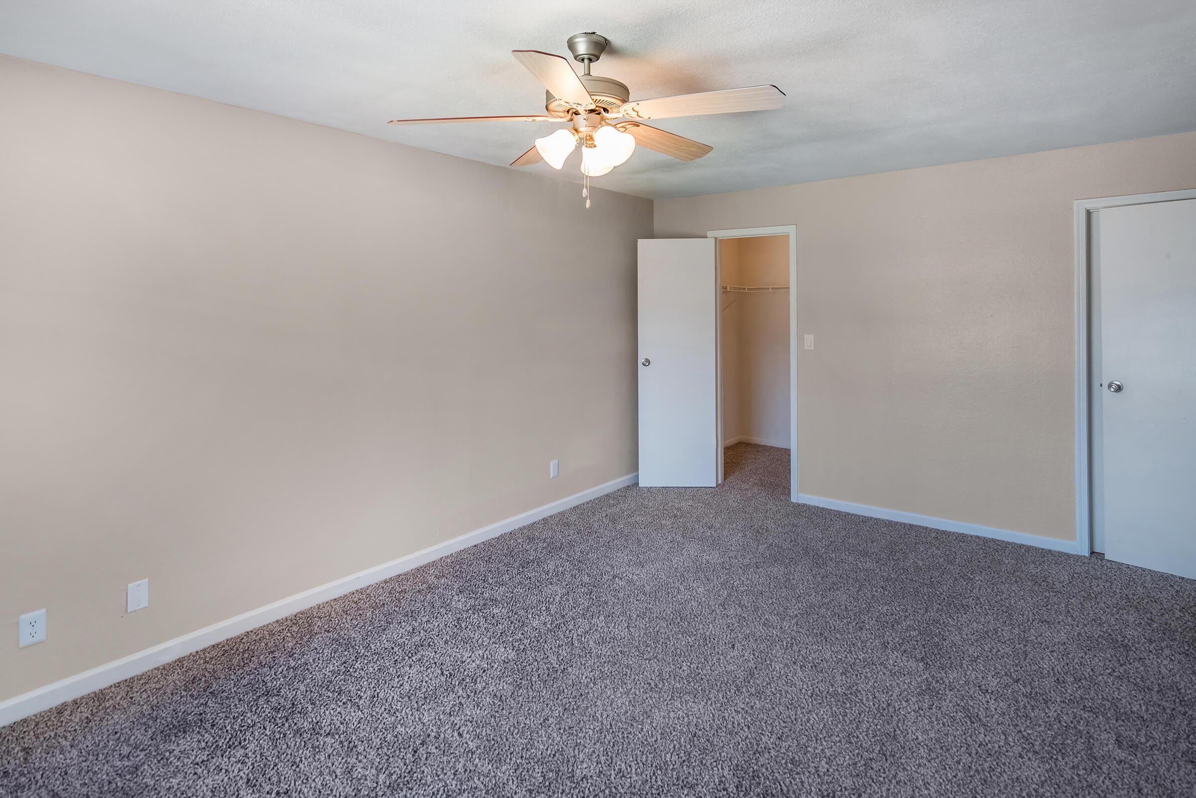 An empty room with light beige walls and a ceiling fan. The floor is covered in gray carpet. A closed door is visible on the right, while an open closet with no door is on the left. Natural light illuminates the space, creating a bright and airy atmosphere.
