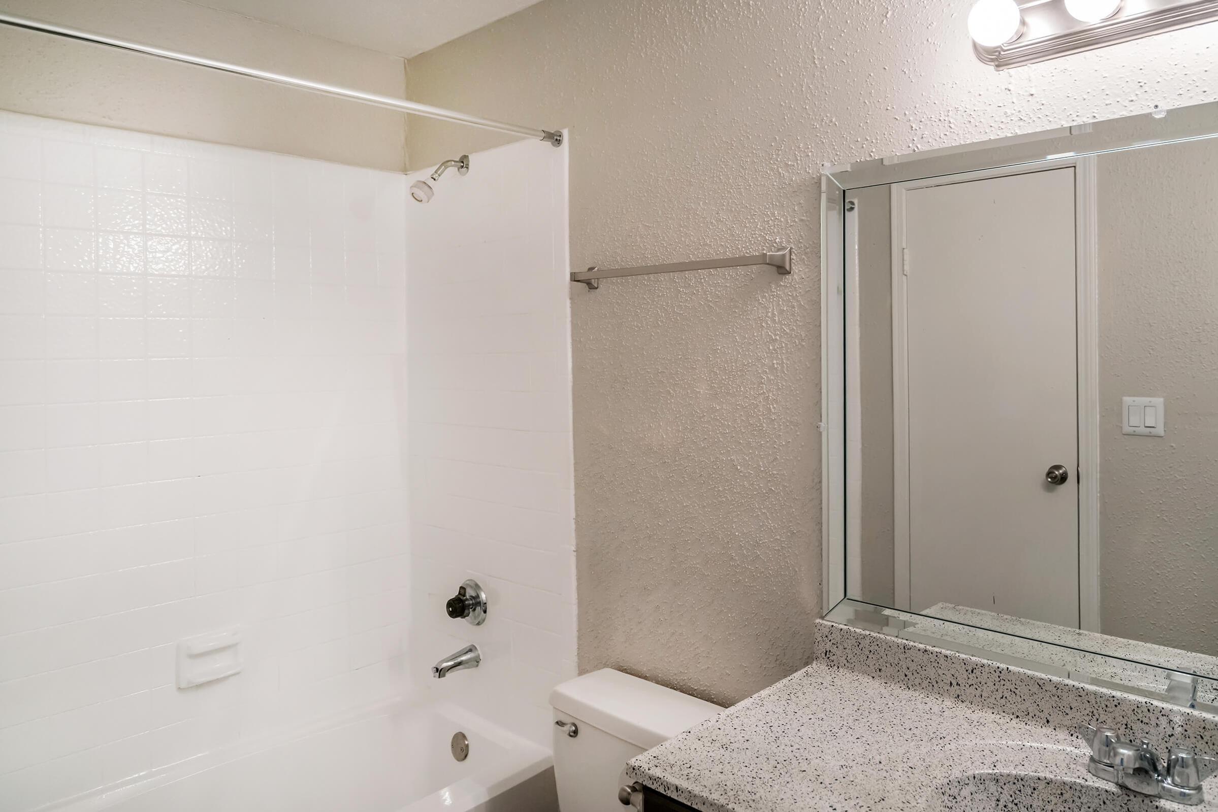 A clean and modern bathroom featuring a white bathtub with a shower, a marble countertop sink, and a square mirror. The walls are painted a light color, and there are simple fixtures and a towel rack visible. The space is well-lit with overhead lighting.