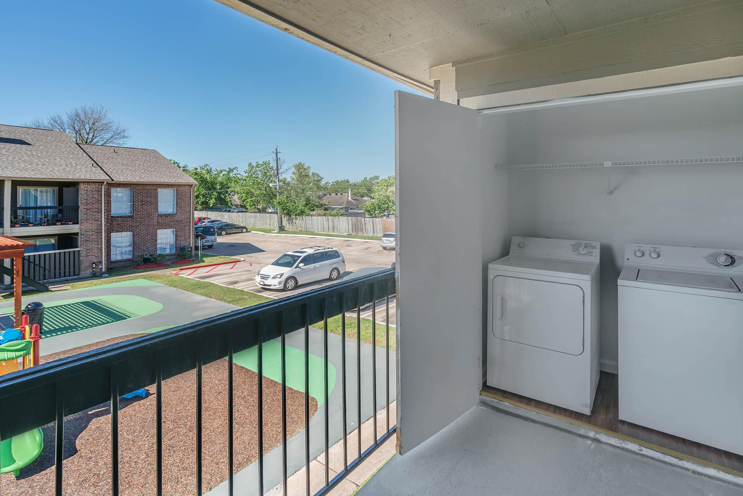 View from a balcony showing a laundry area with a washer and dryer. In the background, there's an apartment community with a playground and green space visible. The sky is clear and blue, indicating a sunny day. A white car is parked nearby on the asphalt.