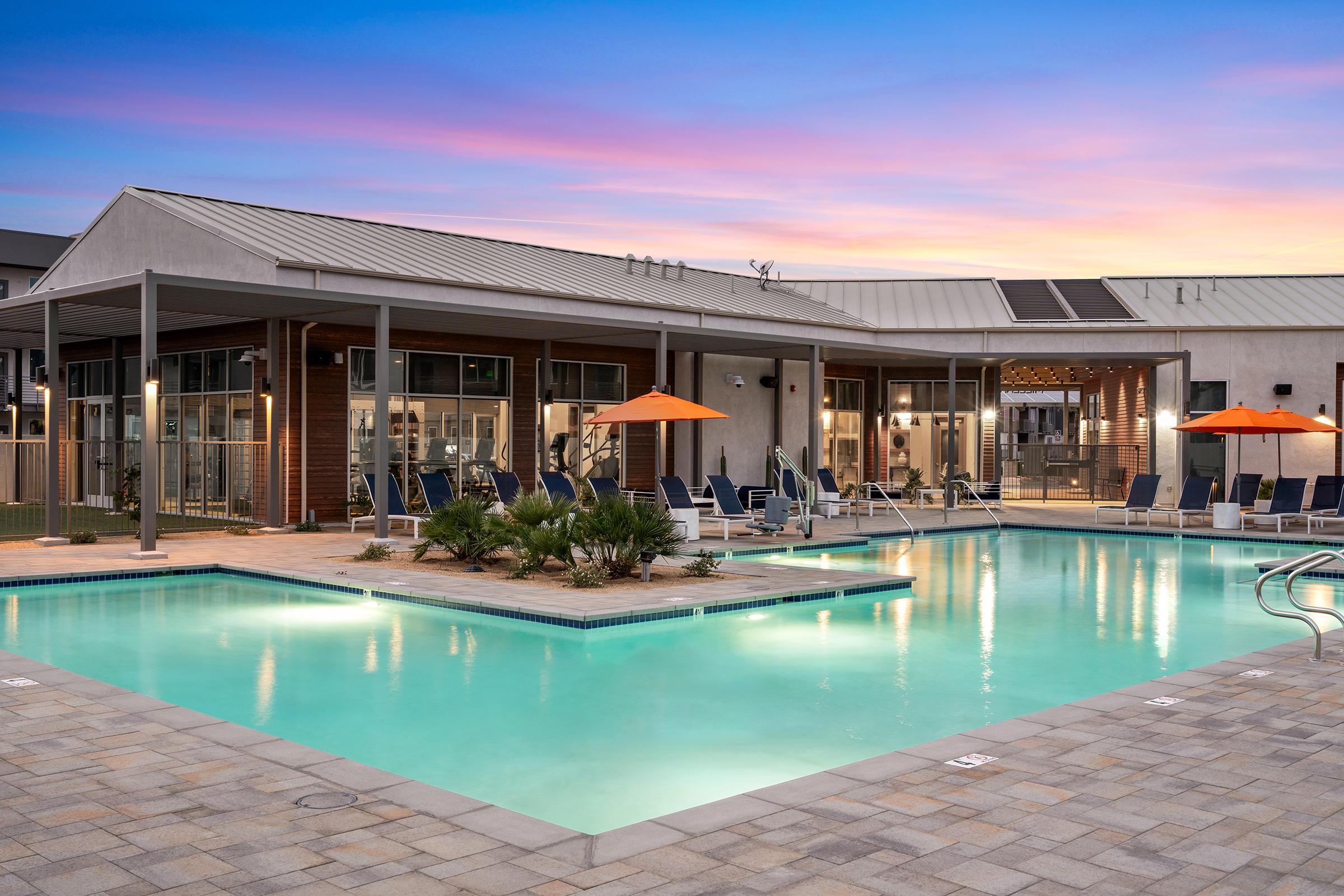 A modern swimming pool surrounded by lounge chairs and umbrellas, with a contemporary building featuring large windows in the background. The sky is painted in soft hues of pink and blue, indicating sunset, creating a relaxing atmosphere.