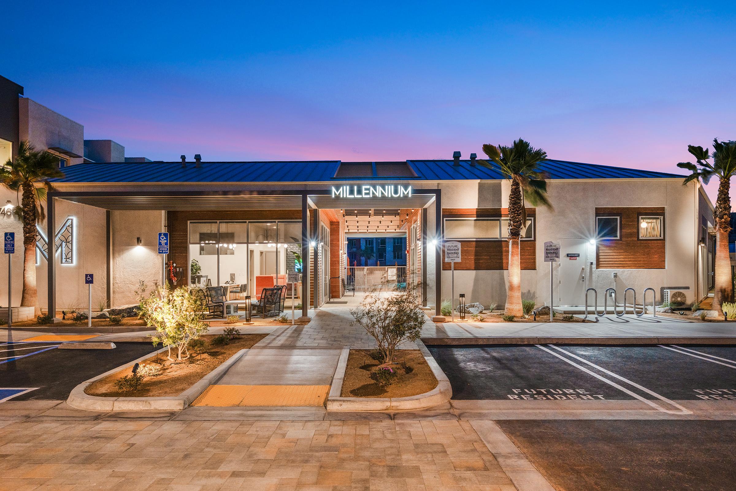 Modern building with the sign "MILLENNIUM" at the entrance, palm trees lining the walkway, and a clear evening sky in the background. The property features a spacious parking area with designated spots and a stylish exterior design, creating an inviting atmosphere.