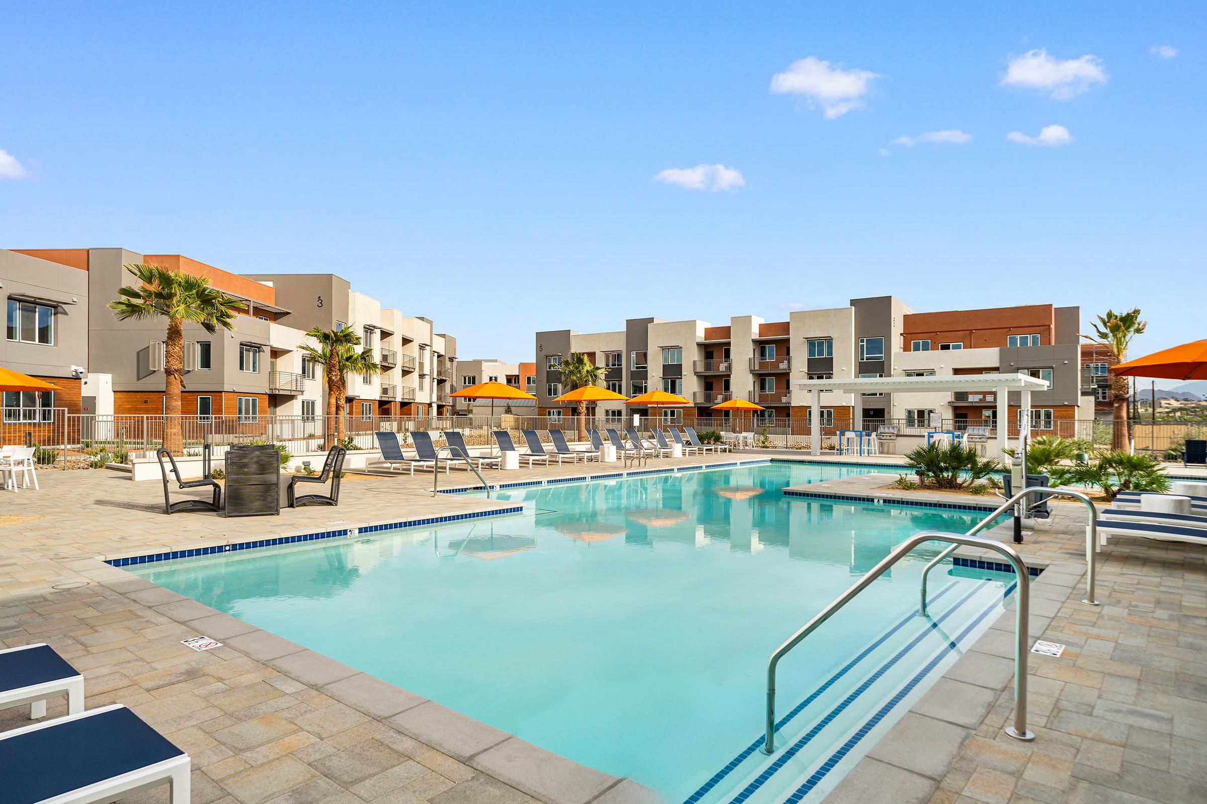 An inviting outdoor pool area surrounded by modern apartment buildings. The pool is surrounded by lounge chairs and umbrellas, with palm trees adding a tropical touch. Clear blue skies overhead create a bright and sunny atmosphere, ideal for relaxation.
