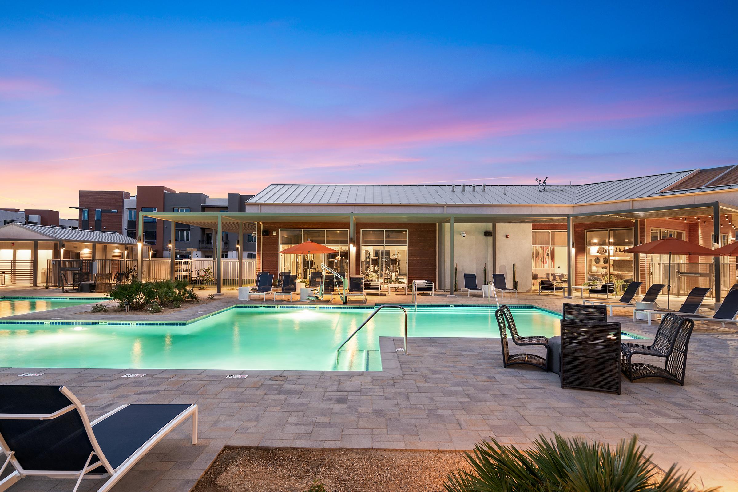 A modern outdoor pool area with a lounge space featuring umbrellas and seating. Surrounding the pool are sleek lounge chairs and a landscaped terrace. The backdrop includes a contemporary building with large windows, under a vibrant sunset sky.