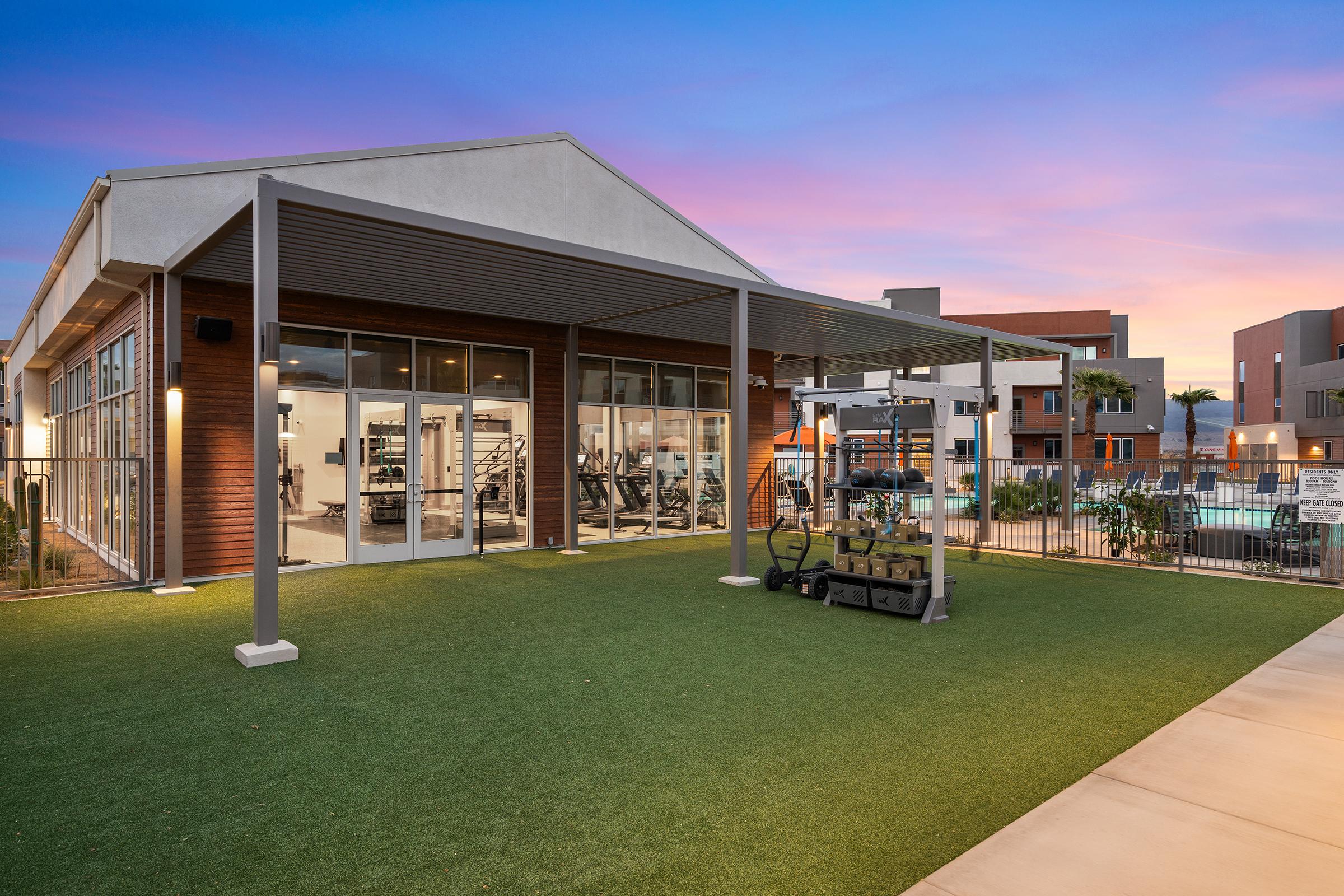 A modern gym facility with large glass windows and a covered outdoor area. The scene includes a lush green lawn, exercise equipment neatly arranged under a shelter, and a pool visible in the background. The sky is painted with soft hues of pink and blue as the sun sets, creating a serene atmosphere.