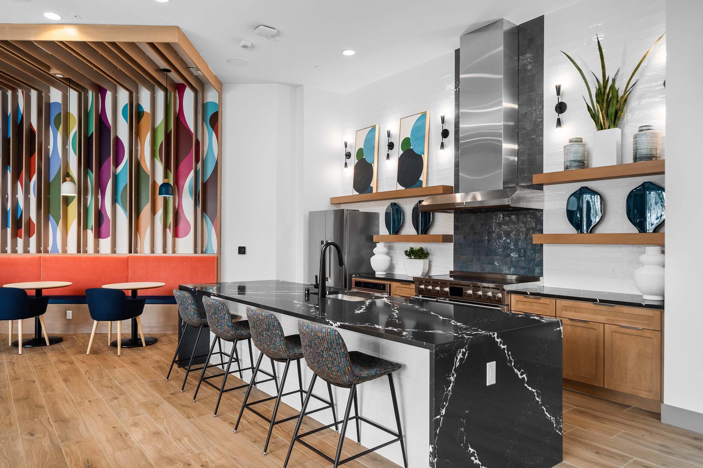 Modern kitchen featuring a large marble island with seating, sleek cabinetry, and stainless steel appliances. The backdrop includes colorful wall art and a stylish wood panel design. Two small dining tables with blue chairs are visible, creating a warm and inviting atmosphere.