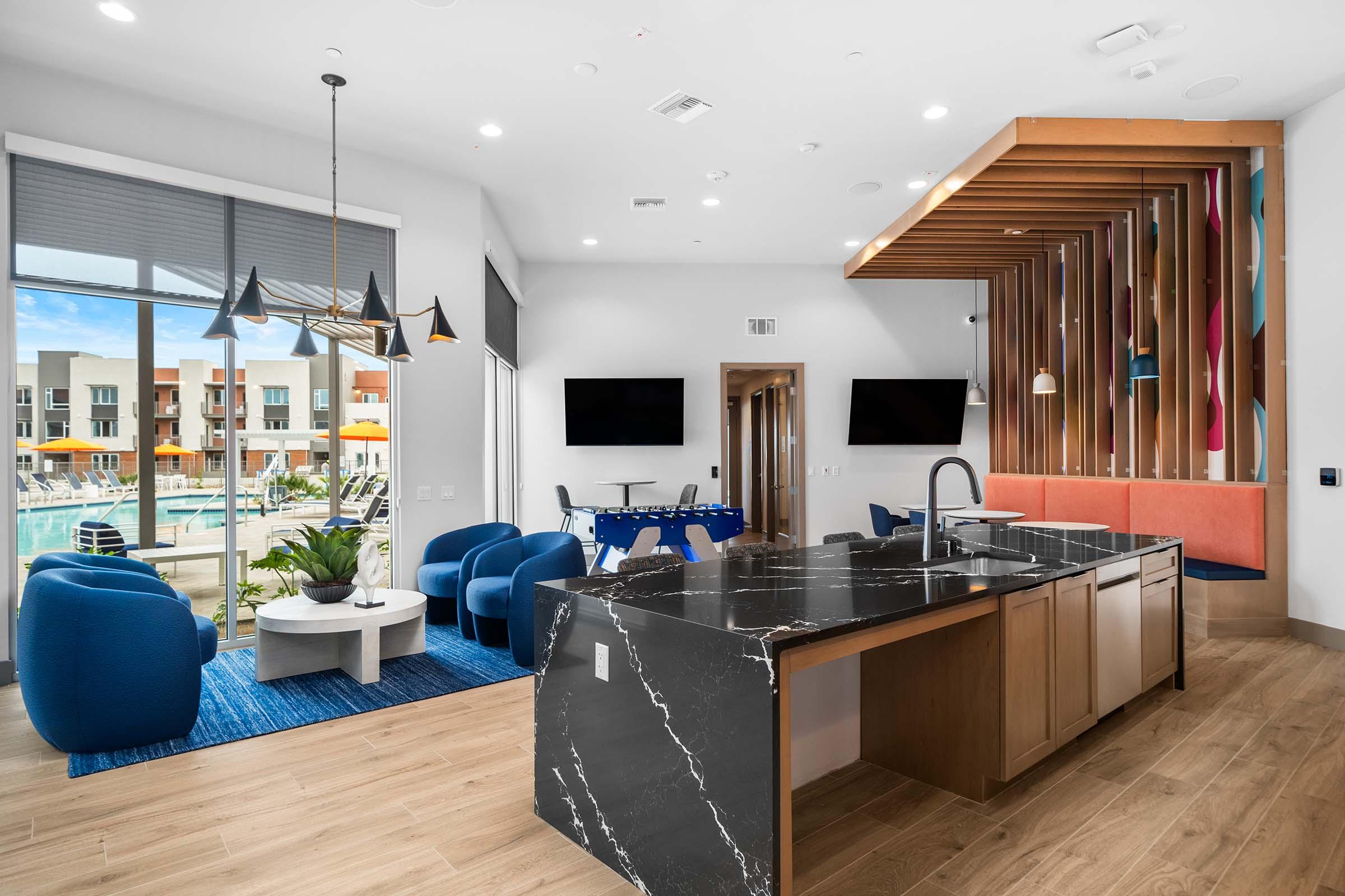 Modern apartment lounge area featuring a black marble kitchen island, blue upholstered chairs, and a round table. Large windows overlook a pool area with outdoor seating. The interior design includes wood accents and contemporary lighting fixtures, creating a welcoming and stylish atmosphere.