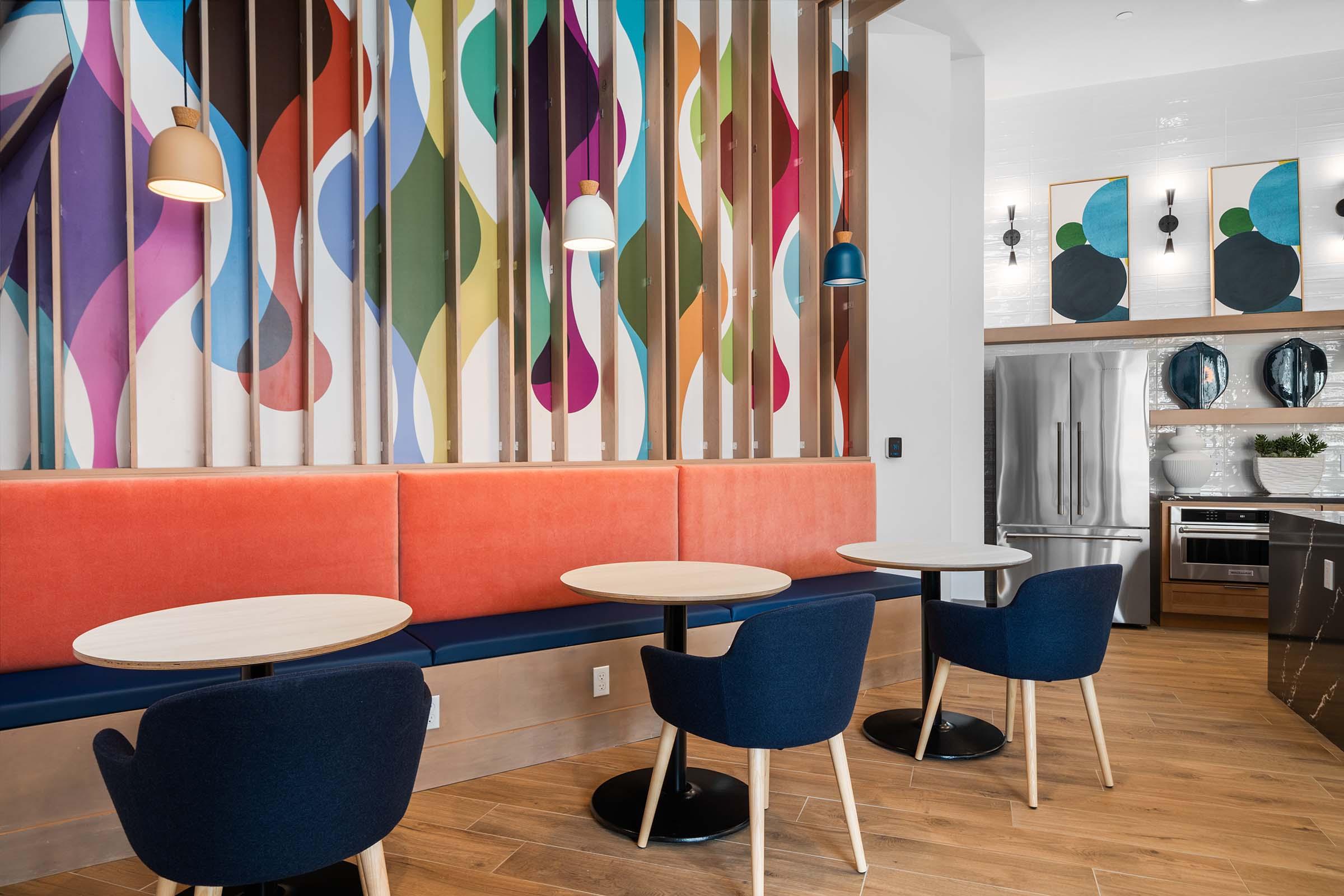 A modern café interior featuring a colorful mural with flowing shapes on a feature wall. There are two round tables with dark blue chairs arranged in front of a bright orange booth. The space is well-lit with pendant lights hanging above and sleek appliances visible in the background.