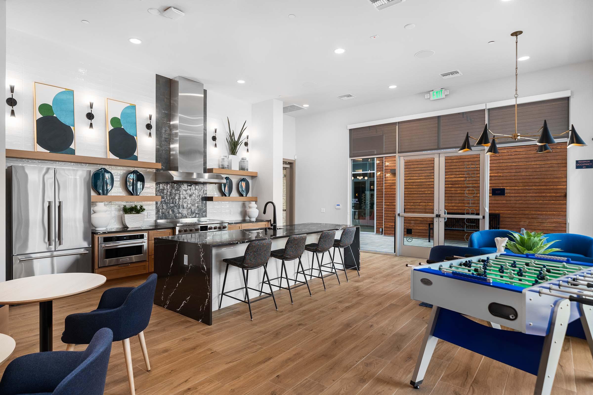 Modern kitchen and lounge area with sleek appliances, a large island with bar seating, and stylish decor. A foosball table is visible in the foreground, while large windows provide natural light. The space features wooden flooring and contemporary furniture in blue tones, creating a welcoming atmosphere for social gatherings.