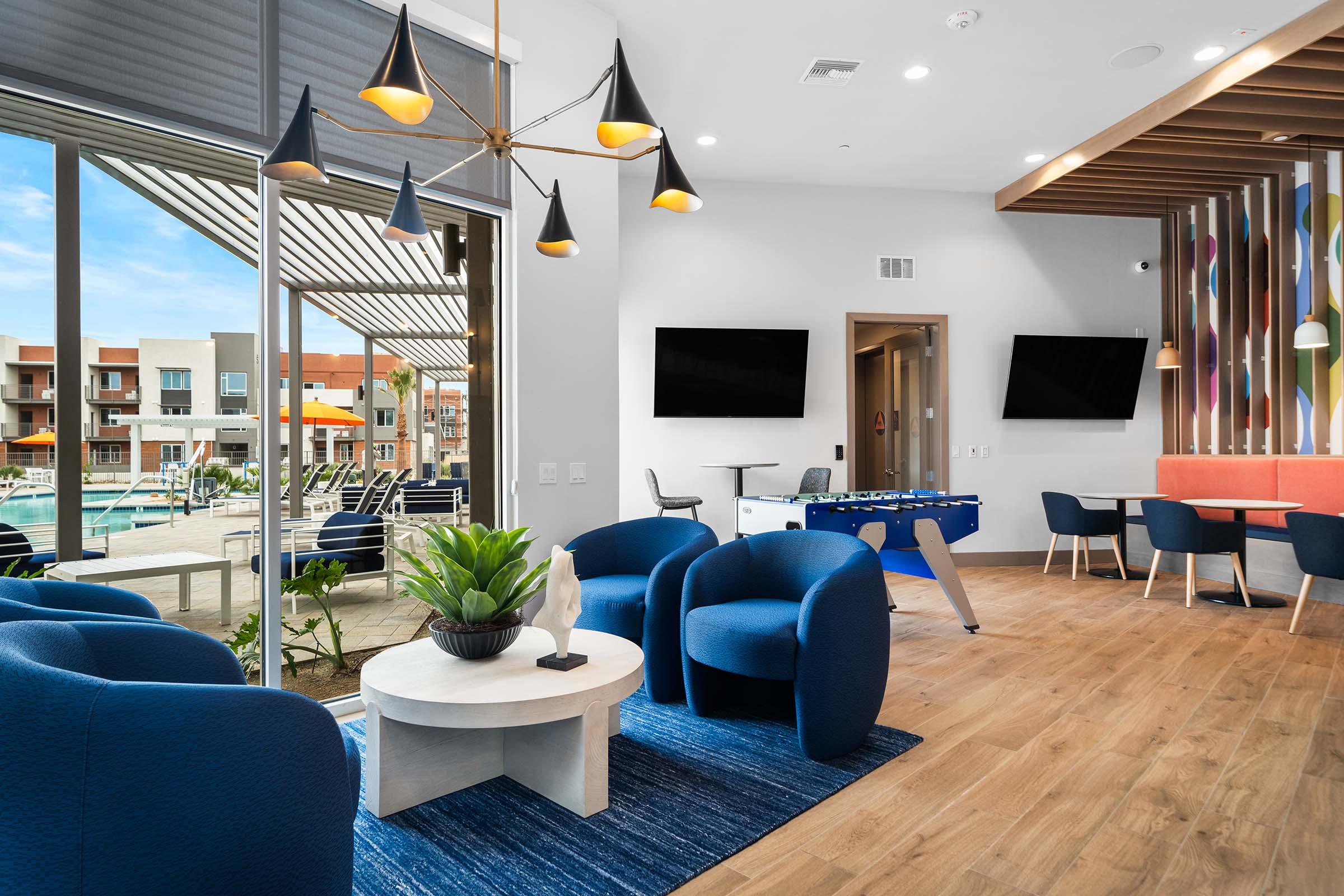 A modern lounge area featuring stylish blue chairs and a round table with a decorative plant. The space is bright, with large windows overlooking a pool area. There are two flat-screen TVs on the wall, a foosball table, and a cozy seating arrangement with colorful accents, creating a welcoming social atmosphere.