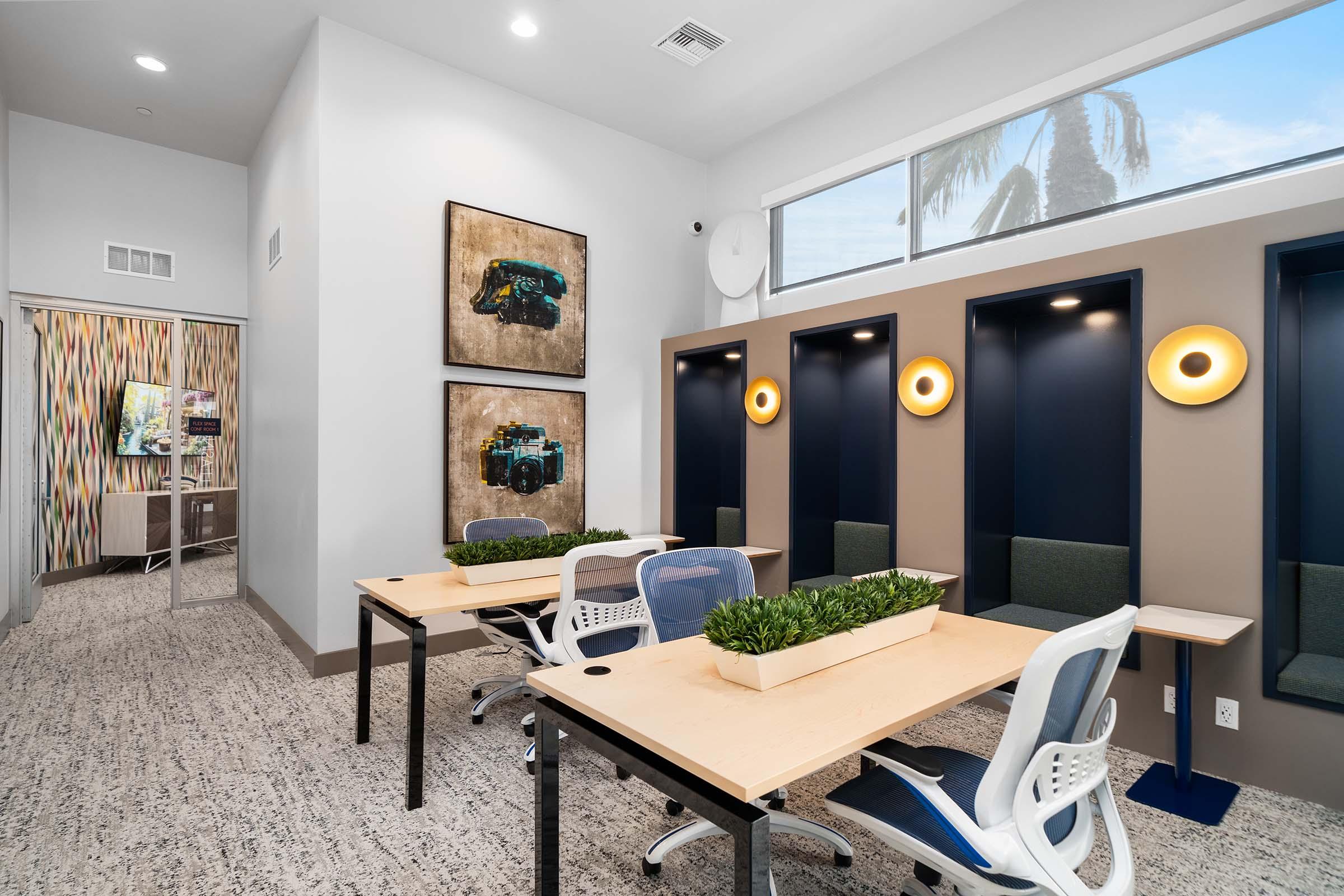 Two modern workstations with light wooden desks and ergonomic chairs are arranged in a bright room. Each desk features a planter with greenery. The walls have abstract art and decorative lighting. A glass door leads to another room, and large windows let in natural light, creating a spacious atmosphere.