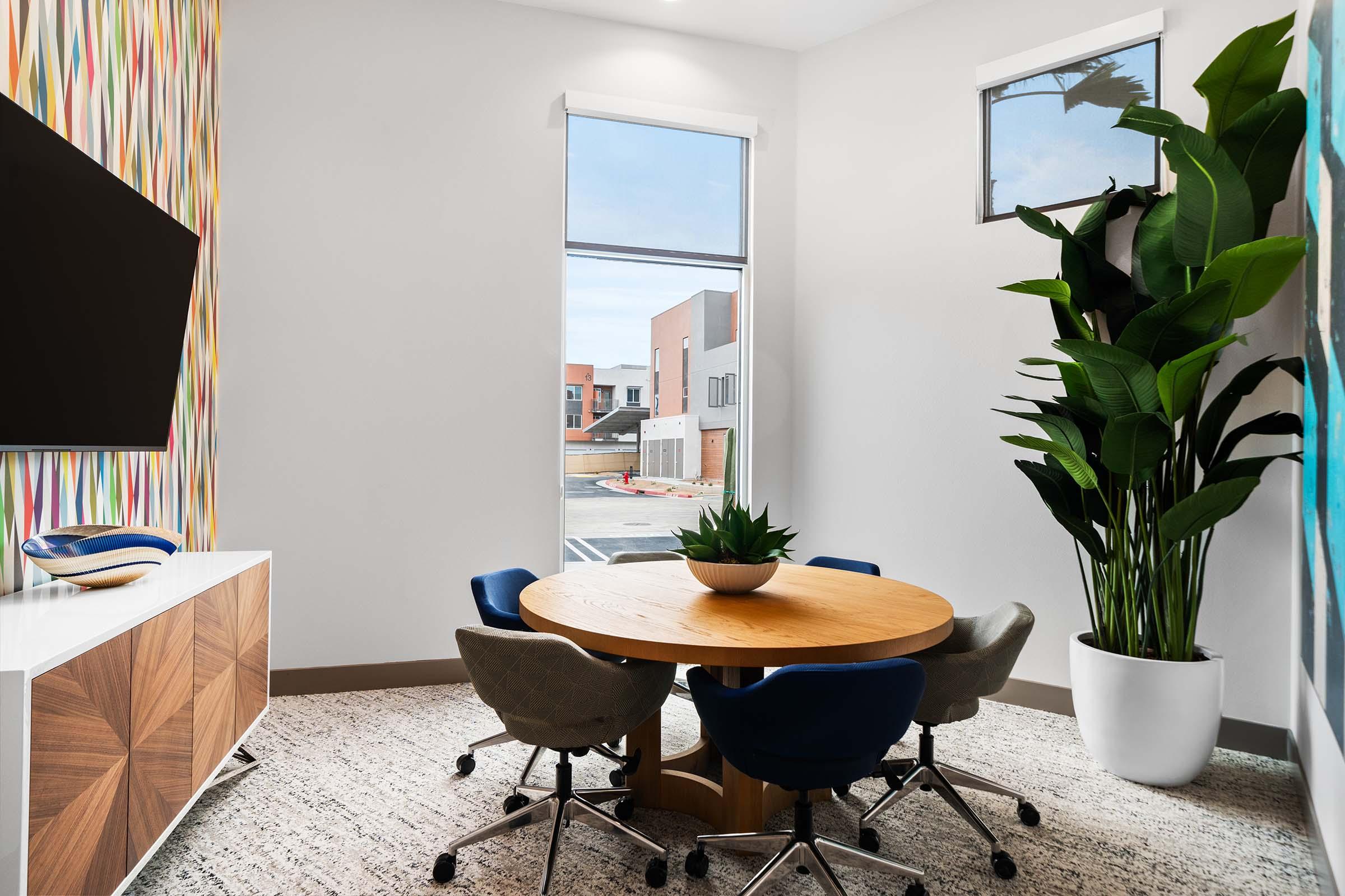 A modern office space featuring a round wooden table surrounded by upholstered chairs. There is a large TV mounted on the wall and a decorative plant in a white pot. The walls showcase colorful geometric patterns, and large windows let in natural light, providing a bright and inviting atmosphere.