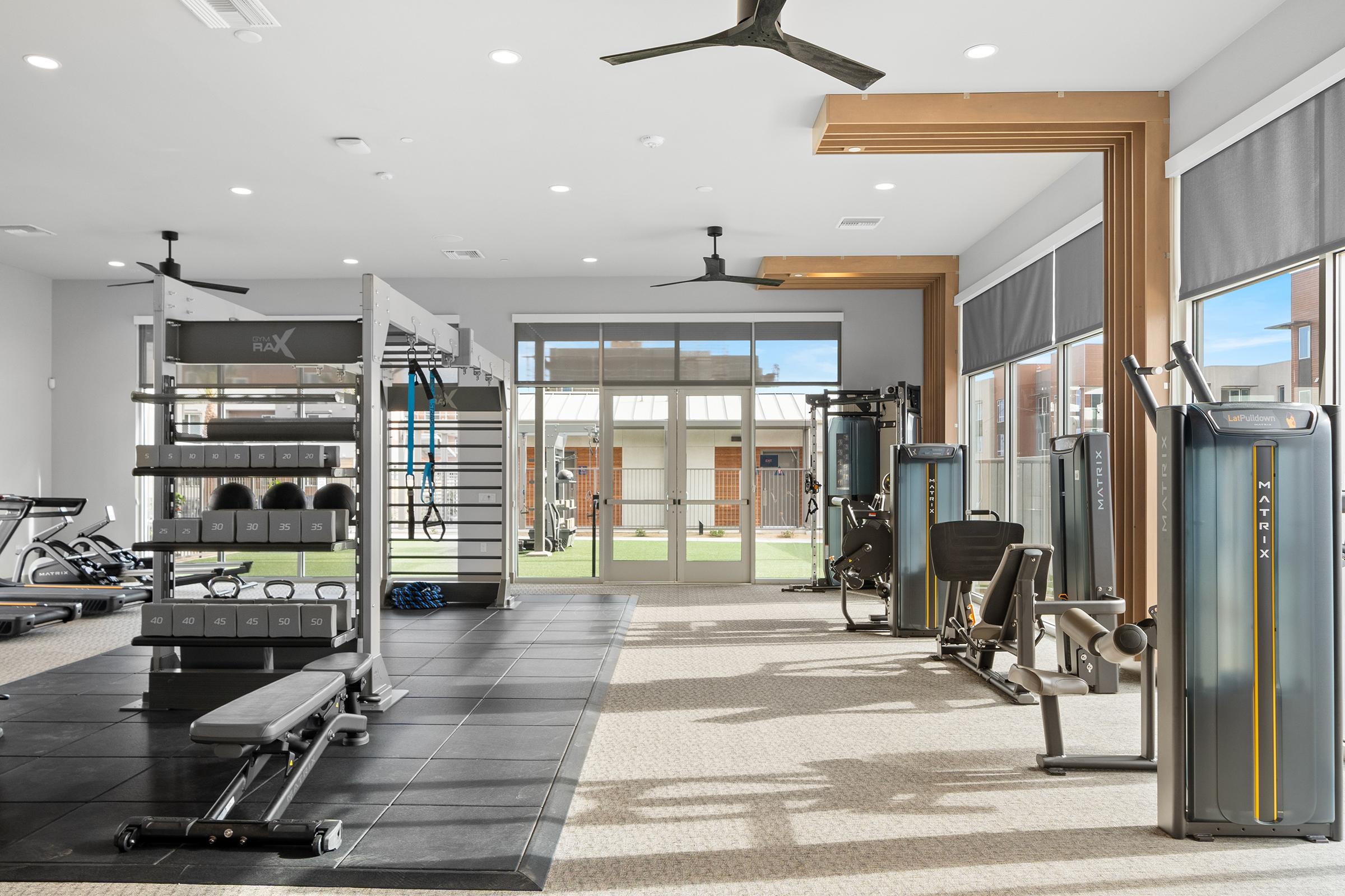 A modern gym interior featuring various exercise equipment, including weight machines and free weights. Large windows provide natural light, and there are ceiling fans for ventilation. The space has a clean, minimalistic design with a mix of dark flooring and light-colored walls.