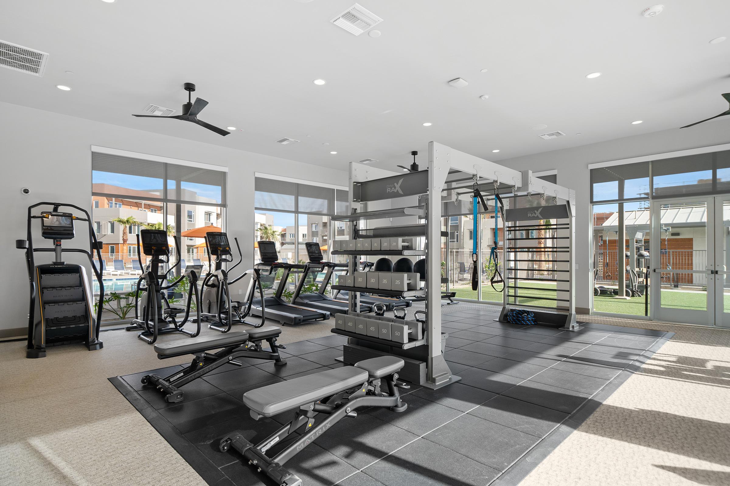 A modern gym interior featuring exercise equipment, including treadmills, stationary bikes, and a multi-station strength training setup. Large windows let in natural light and provide a view of an outdoor pool area. The flooring is covered with black mats, and the space has a bright, inviting atmosphere.