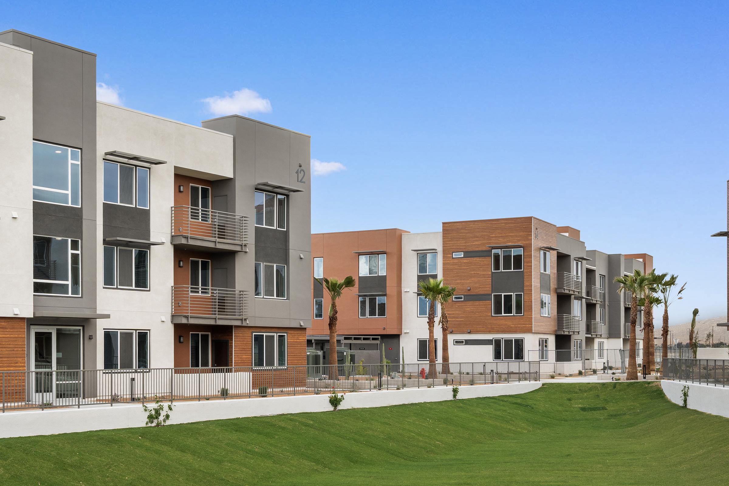Modern apartment units featuring a mix of white, gray, and wooden exteriors. The buildings have balconies and large windows, set against a bright blue sky. A manicured green lawn with palm trees runs in front of the complex, providing a welcoming outdoor space.