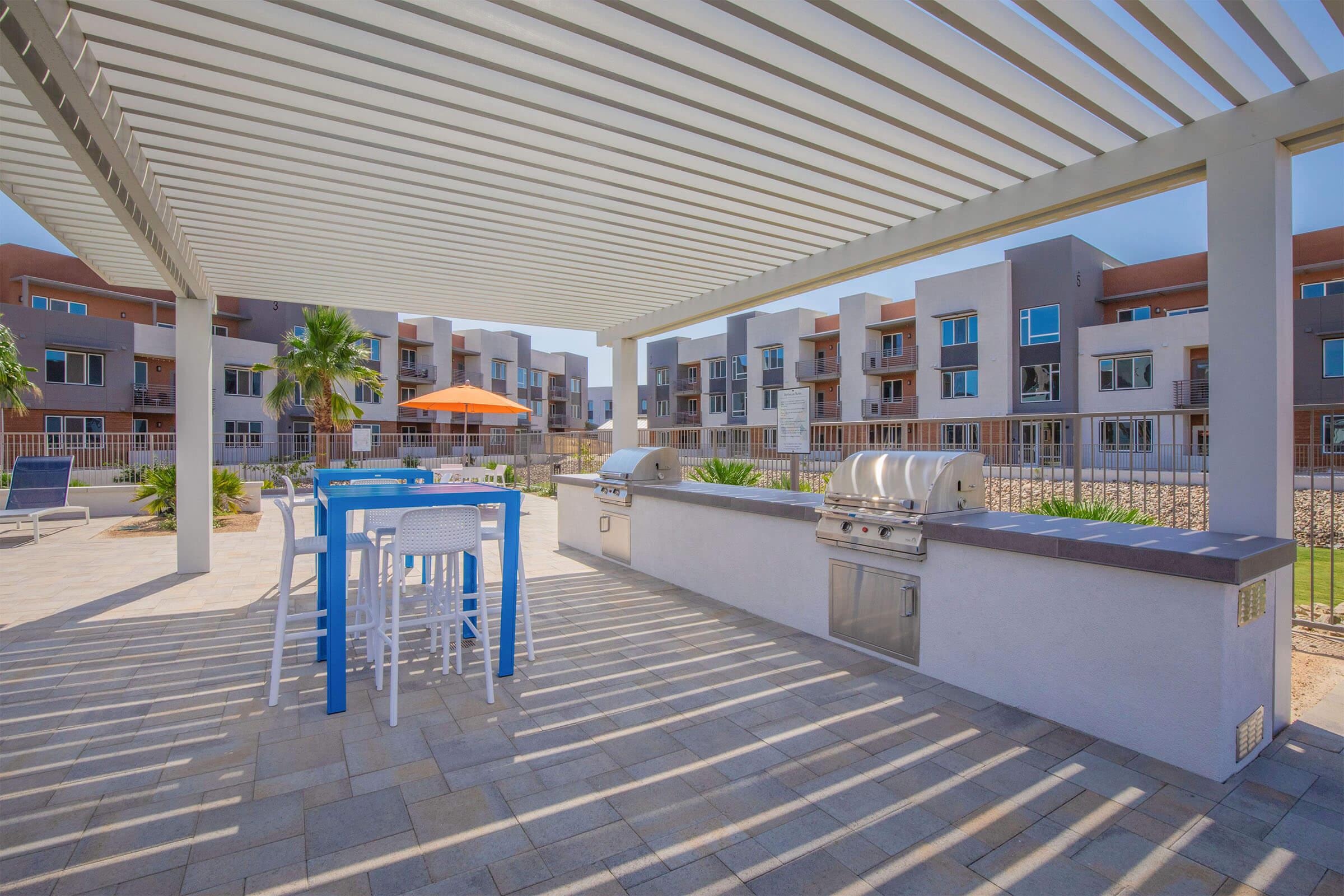 Outdoor barbecue area with a grill and seating, shaded by a pergola. In the background, modern apartment buildings are visible. The scene is sunny, with an orange umbrella providing additional shade. The space features pavers and landscaping, creating a welcoming atmosphere for gatherings.