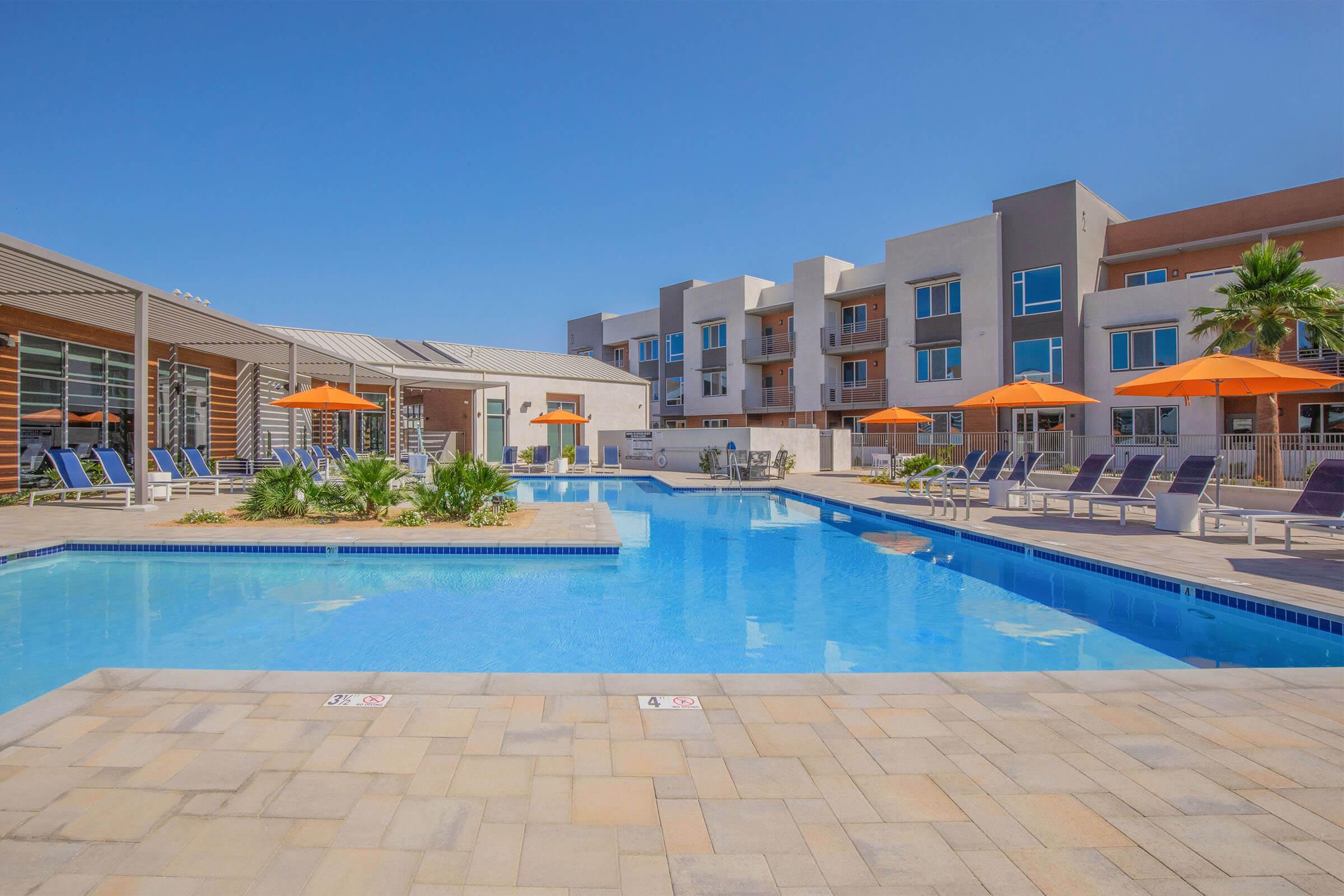 A bright, sunny pool area featuring a large, clear blue swimming pool surrounded by lounge chairs and orange umbrellas. Modern buildings are visible in the background, with palm trees adding to the relaxed, tropical atmosphere. The scene is inviting and perfect for leisure activities.