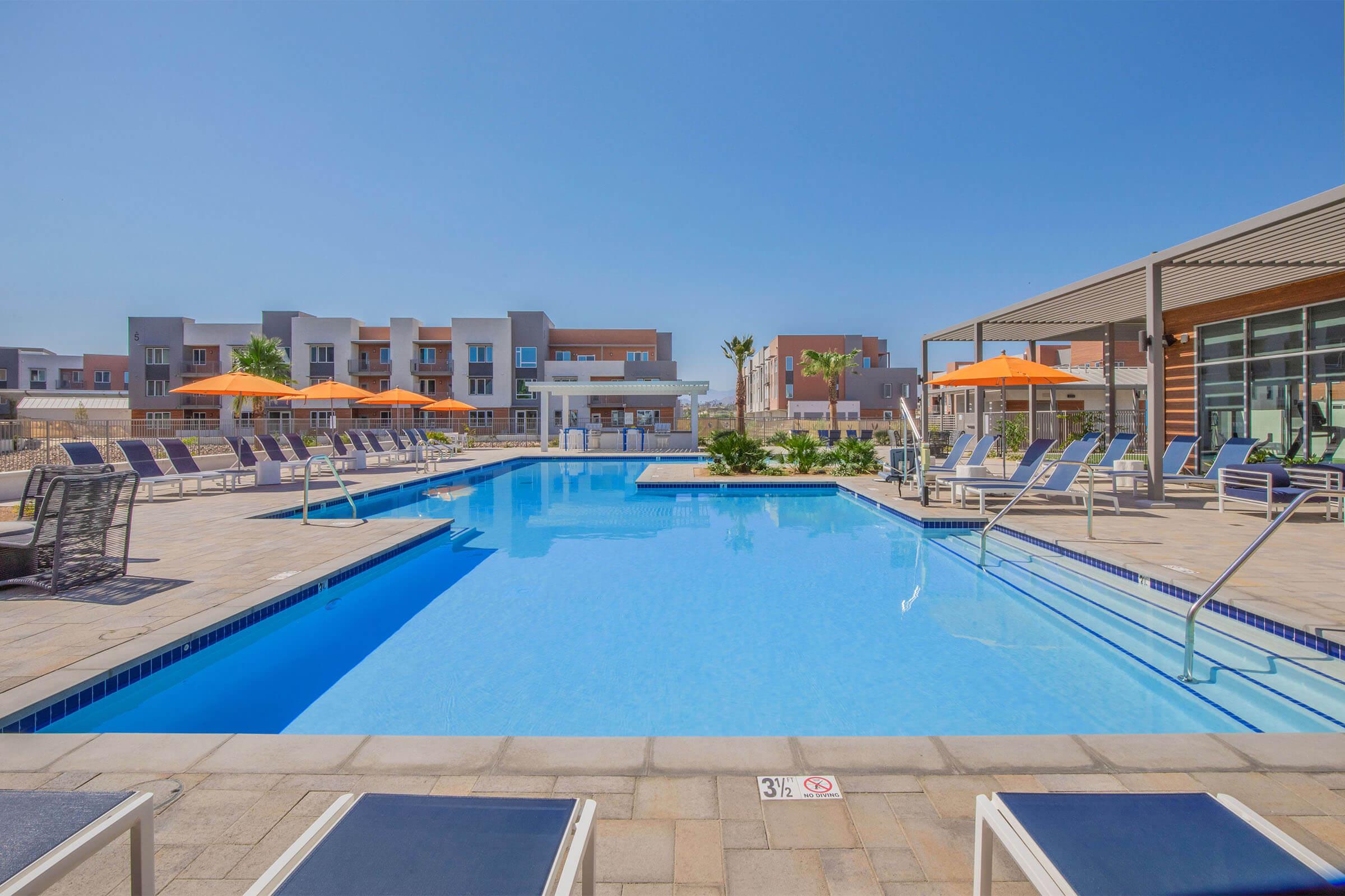 A bright and inviting pool area featuring a clear blue swimming pool surrounded by lounge chairs and vibrant orange umbrellas. In the background, modern buildings and palm trees complete the sunny, relaxing atmosphere, ideal for leisure and enjoyment.
