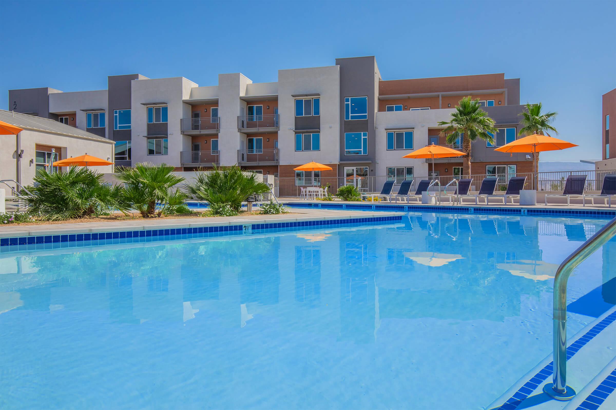 A serene swimming pool with clear blue water, surrounded by lounge chairs and vibrant orange umbrellas. In the background, modern buildings with balconies can be seen, complemented by palm trees, under a clear blue sky. The setting conveys a relaxing vacation atmosphere.