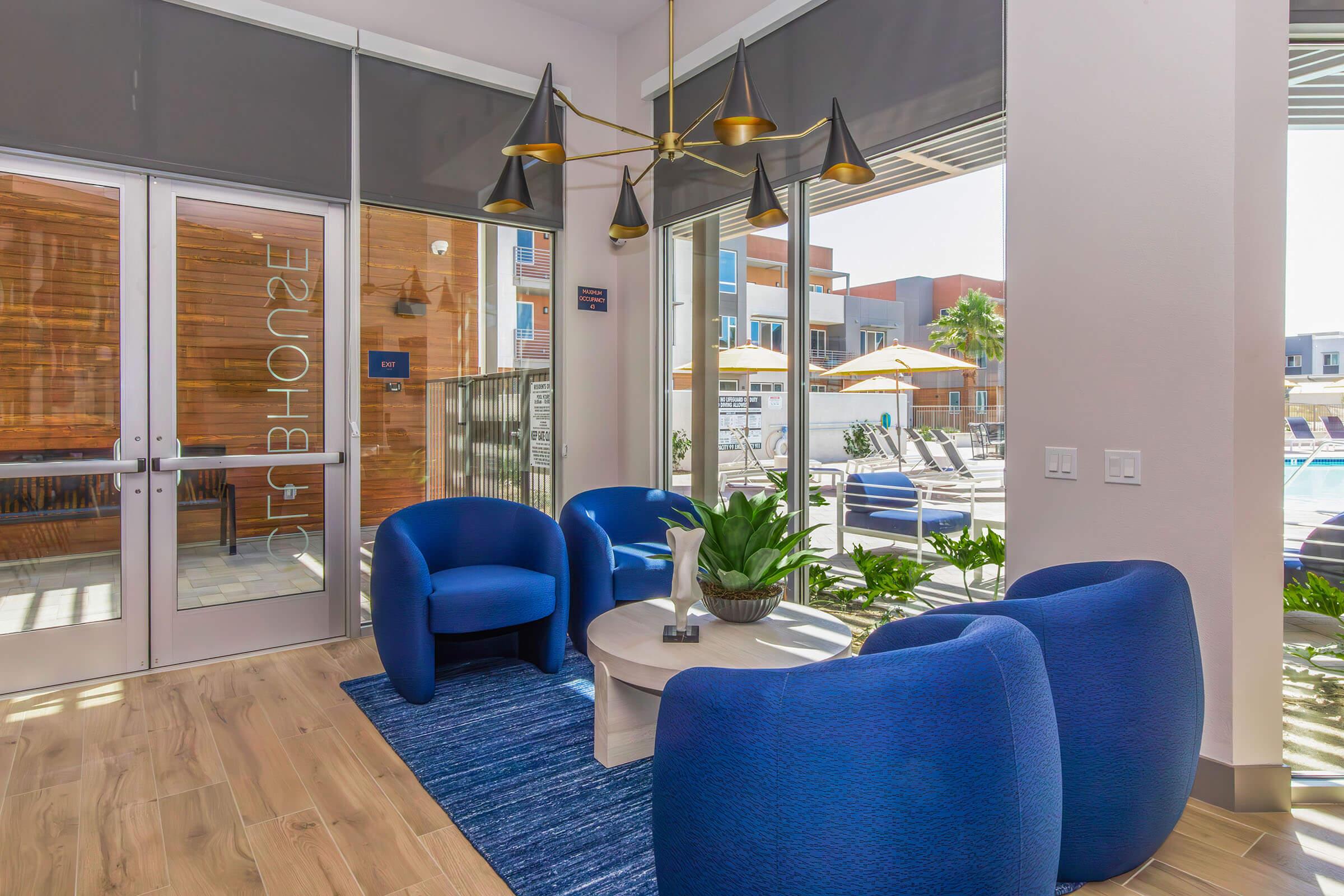 A modern lounge area featuring two blue upholstered chairs and a round coffee table. There's a decorative plant on the table and a stylish light fixture overhead. Large windows provide natural light, with a view of an outdoor pool area and sun loungers. The decor is sleek and contemporary, creating a welcoming atmosphere.