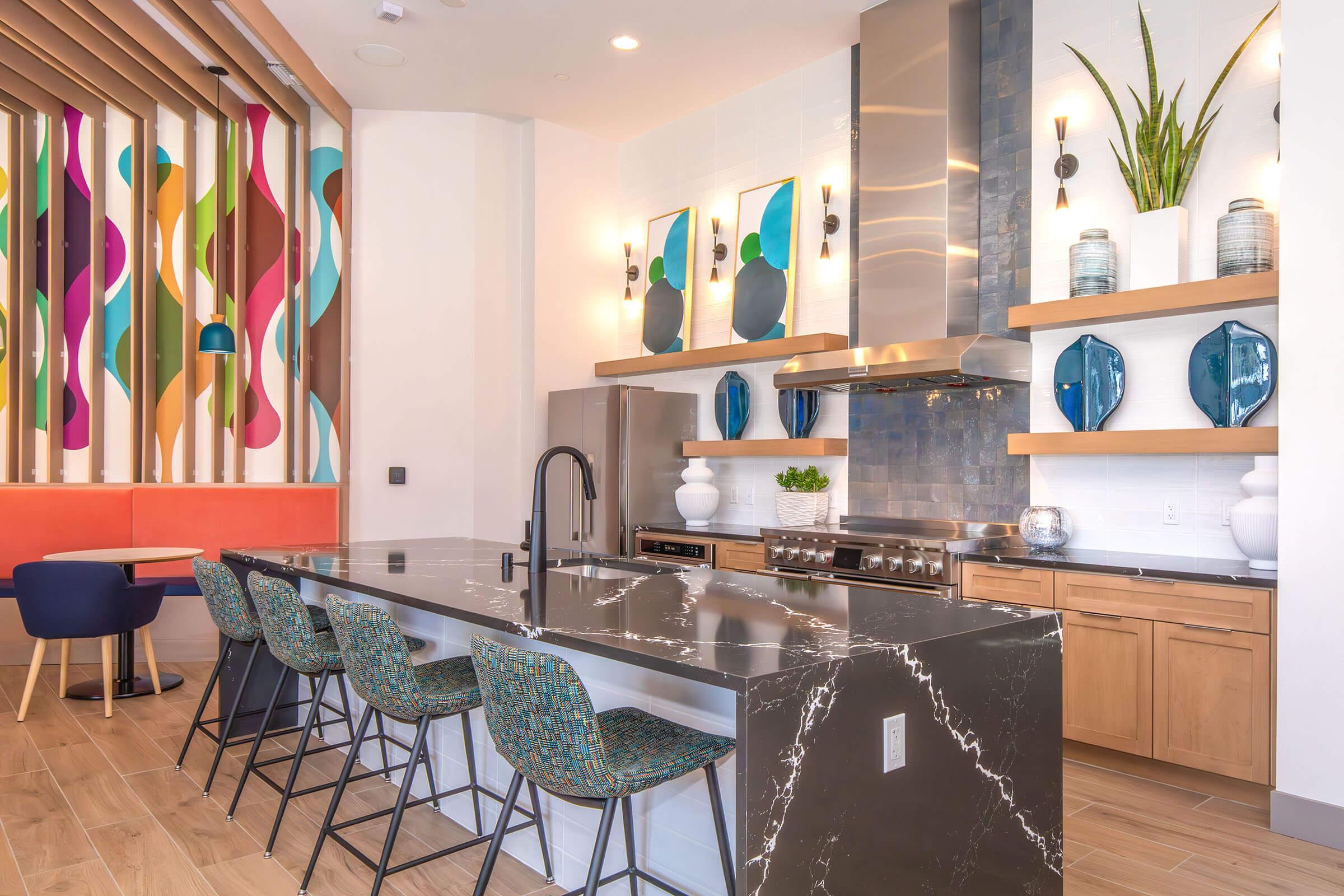 Modern kitchen with a large dark marble island and seating at the counter. Stainless steel appliances, including a refrigerator and oven. Decorative shelves display blue vases and plants. A colorful wall with abstract art adds vibrancy to the space. Light wood flooring completes the contemporary design.