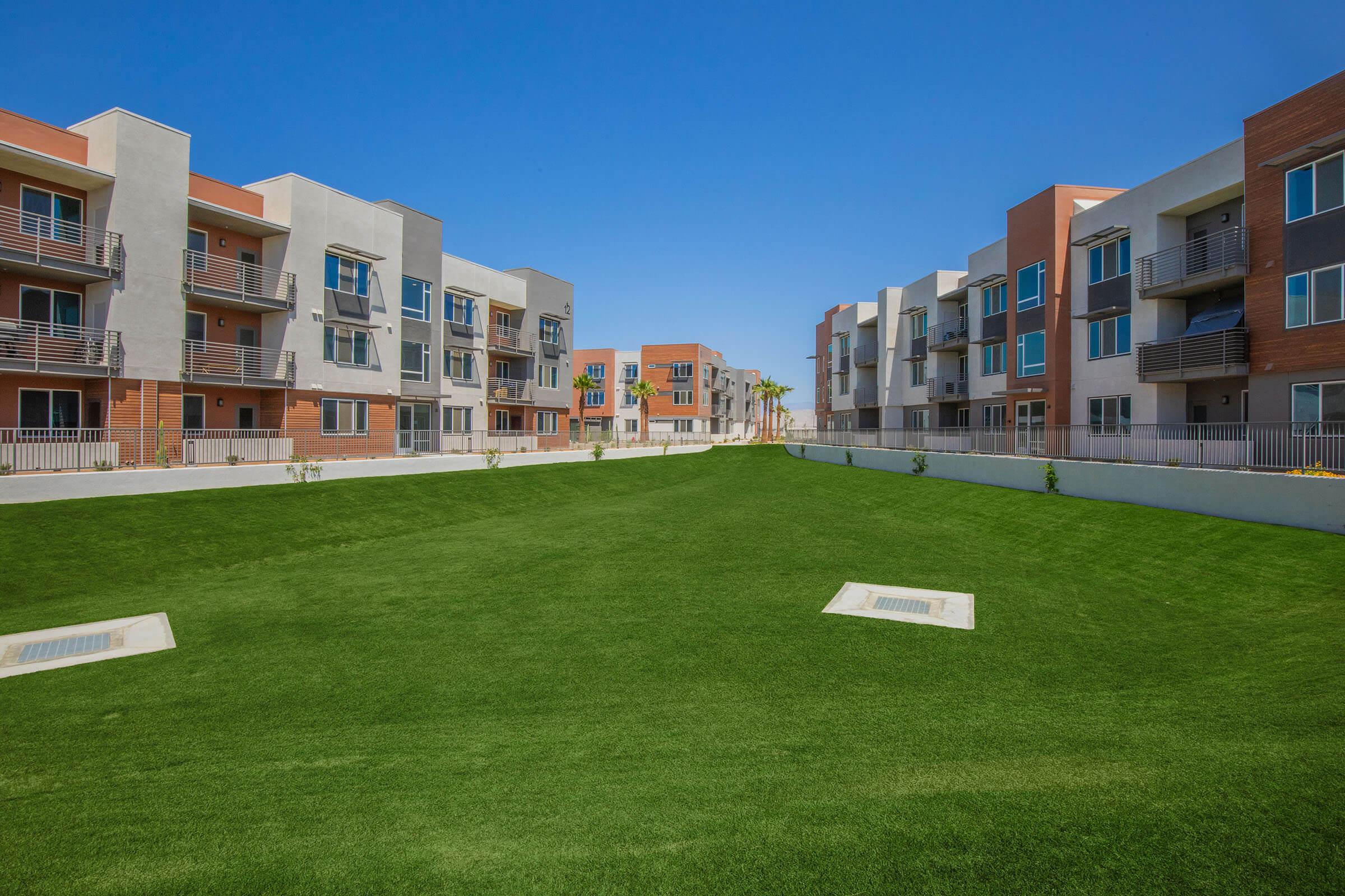 Modern apartment complex with two rows of multi-story buildings facing each other. A spacious green lawn in the center, surrounded by pathways. Clear blue sky above creates a bright and inviting atmosphere.