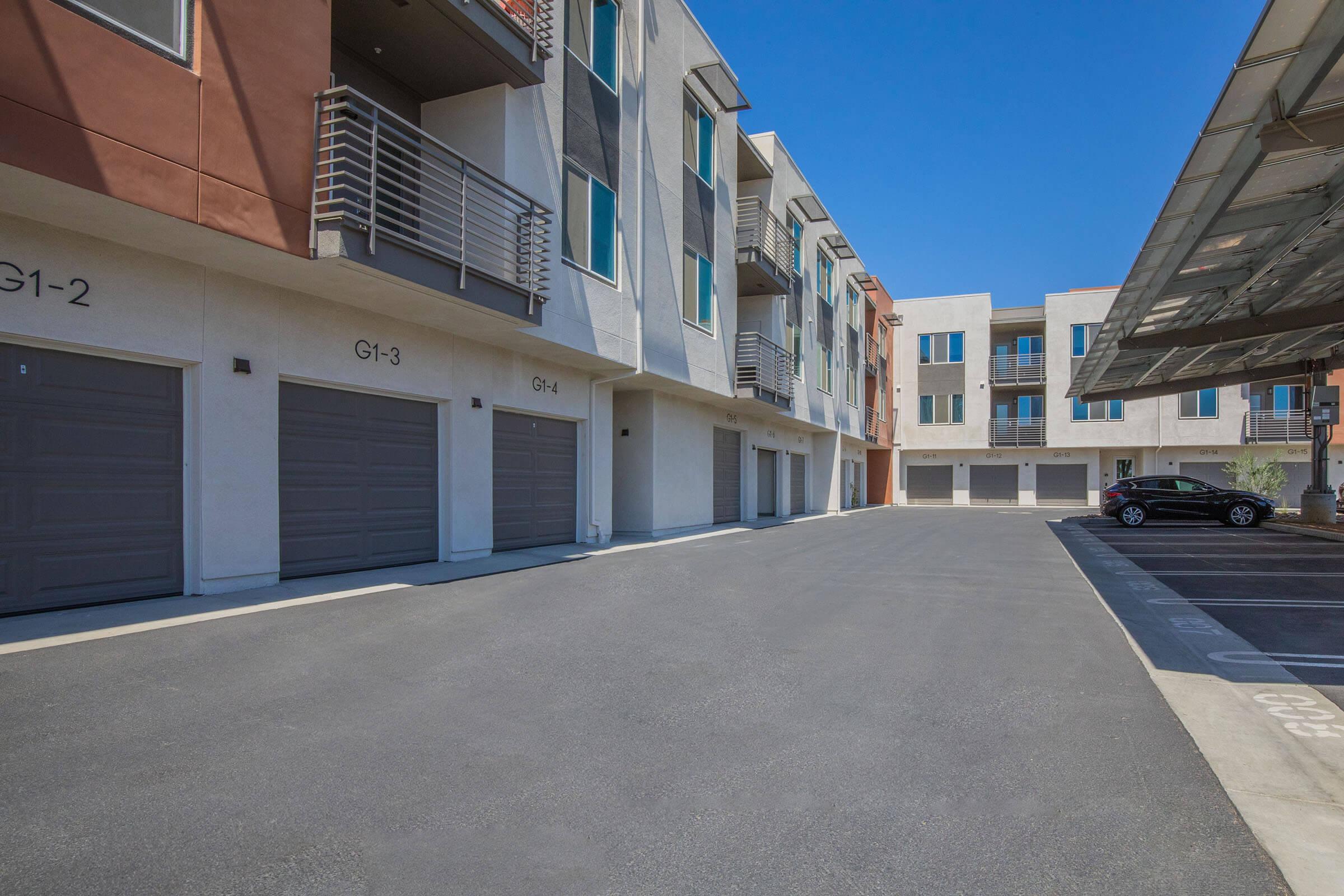 A view of a modern residential building complex featuring several multi-story units with balconies. In the foreground, there is a paved parking area with individual garage doors labeled G1-2, G1-3, and G1-4. A section of the building is shaded by solar panels, and a parked car is visible nearby. The sky is clear and blue.
