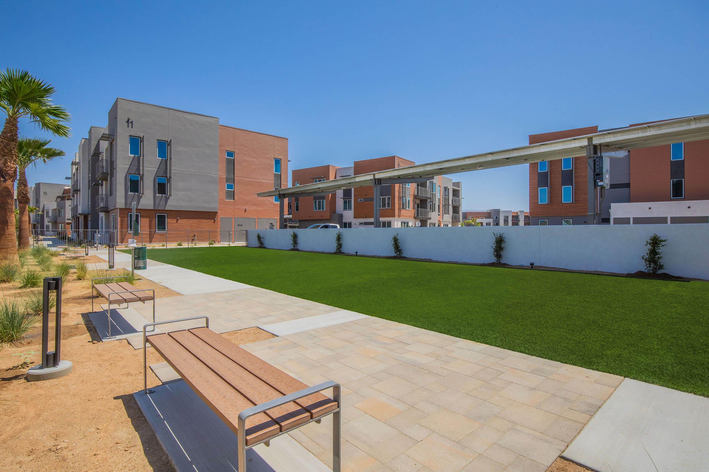 A modern residential area featuring multiple apartment buildings with a clear blue sky. In the foreground, there is a landscaped area with artificial grass, stone walkways, and a bench, creating a communal outdoor space. Palm trees add to the ambiance, enhancing the contemporary setting.