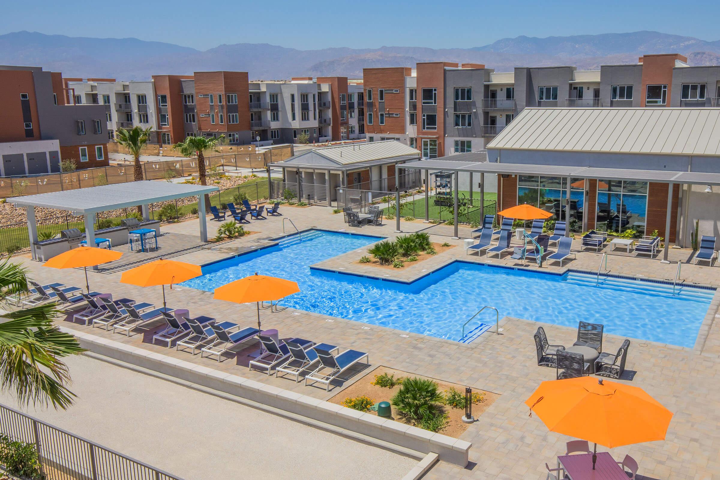 A panoramic view of a modern apartment complex featuring a large swimming pool surrounded by lounge chairs and orange umbrellas. There are shaded seating areas and a clubhouse in the background, with mountains visible on the horizon under a clear blue sky. Lush landscaping enhances the outdoor space.