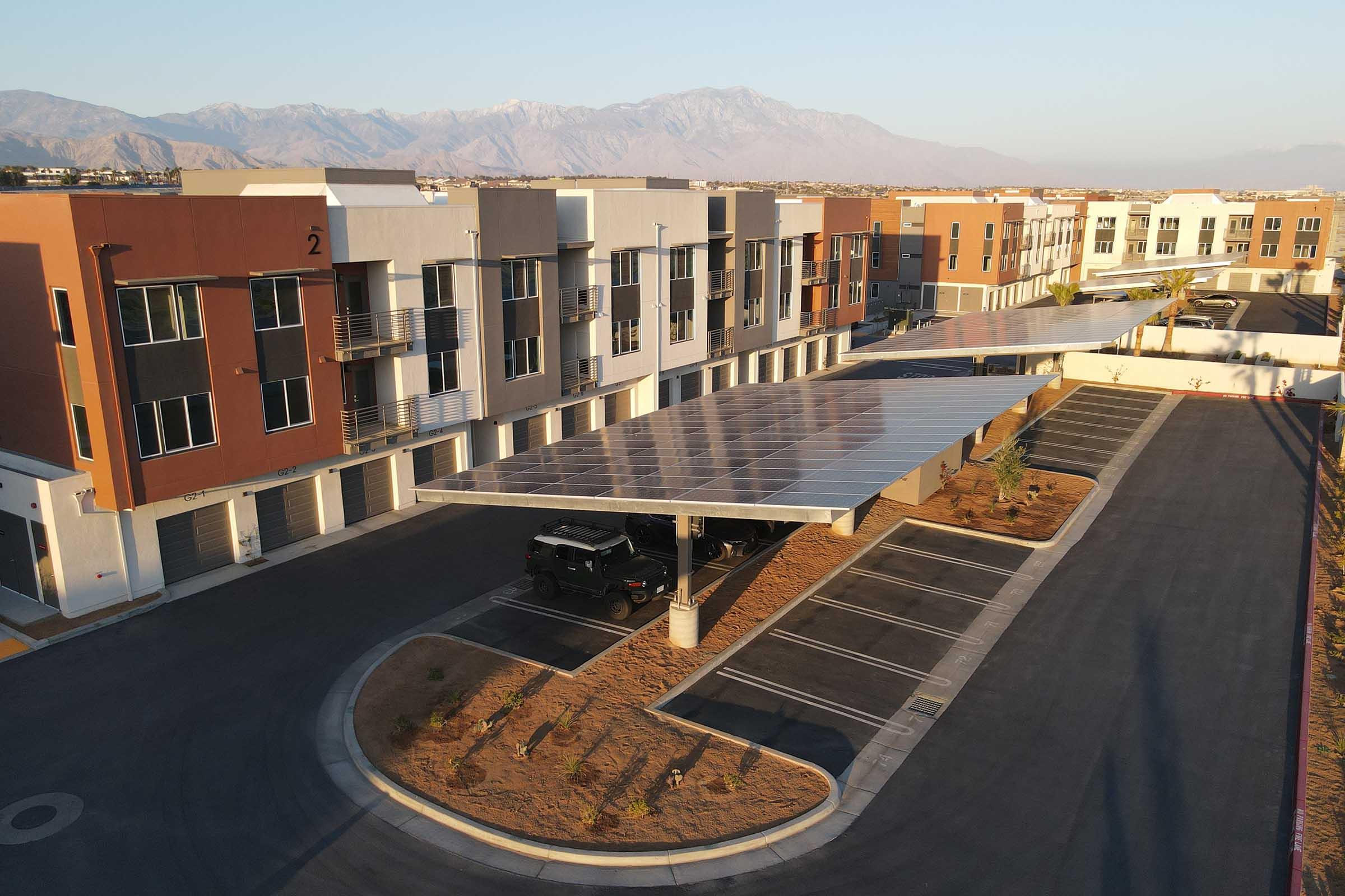 Aerial view of a modern residential complex featuring multi-story buildings with a solar panel-covered parking area. Mountains are visible in the background, and the landscape includes neat landscaping and empty parking spaces in the foreground, highlighting a blend of urban living and sustainability.