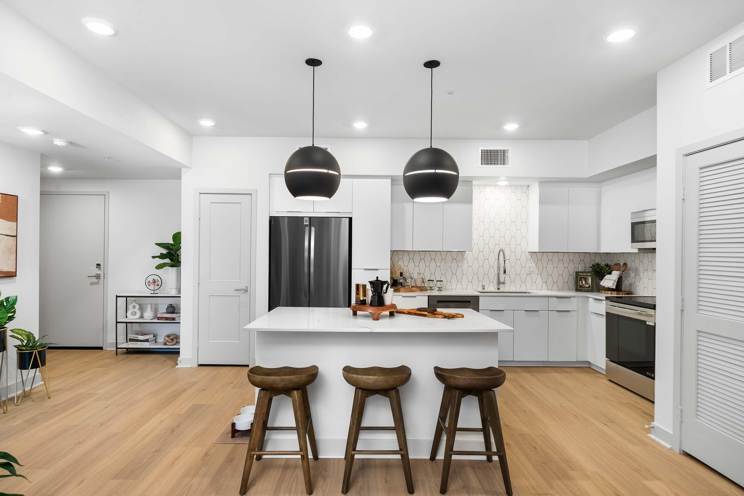 A modern kitchen featuring a central island with three wooden stools, sleek white cabinetry, stainless steel appliances, and geometric tile backsplash. Pendant lights hang above the island, and there are decorative plants and kitchenware visible. The space has a bright and open design with light wood flooring.