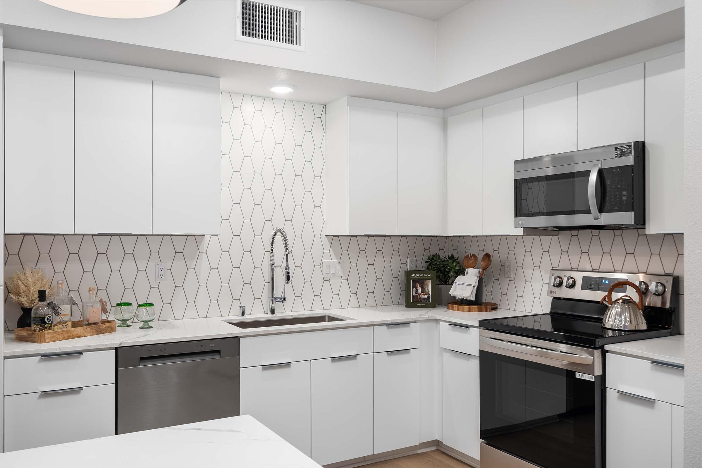 Contemporary kitchen featuring white cabinets, a stainless steel microwave, an electric oven, and a modern backsplash with hexagonal tiles. The sink is set into a spacious countertop with a wooden cutting board and decorative items, complemented by a small plant and green glassware on display.