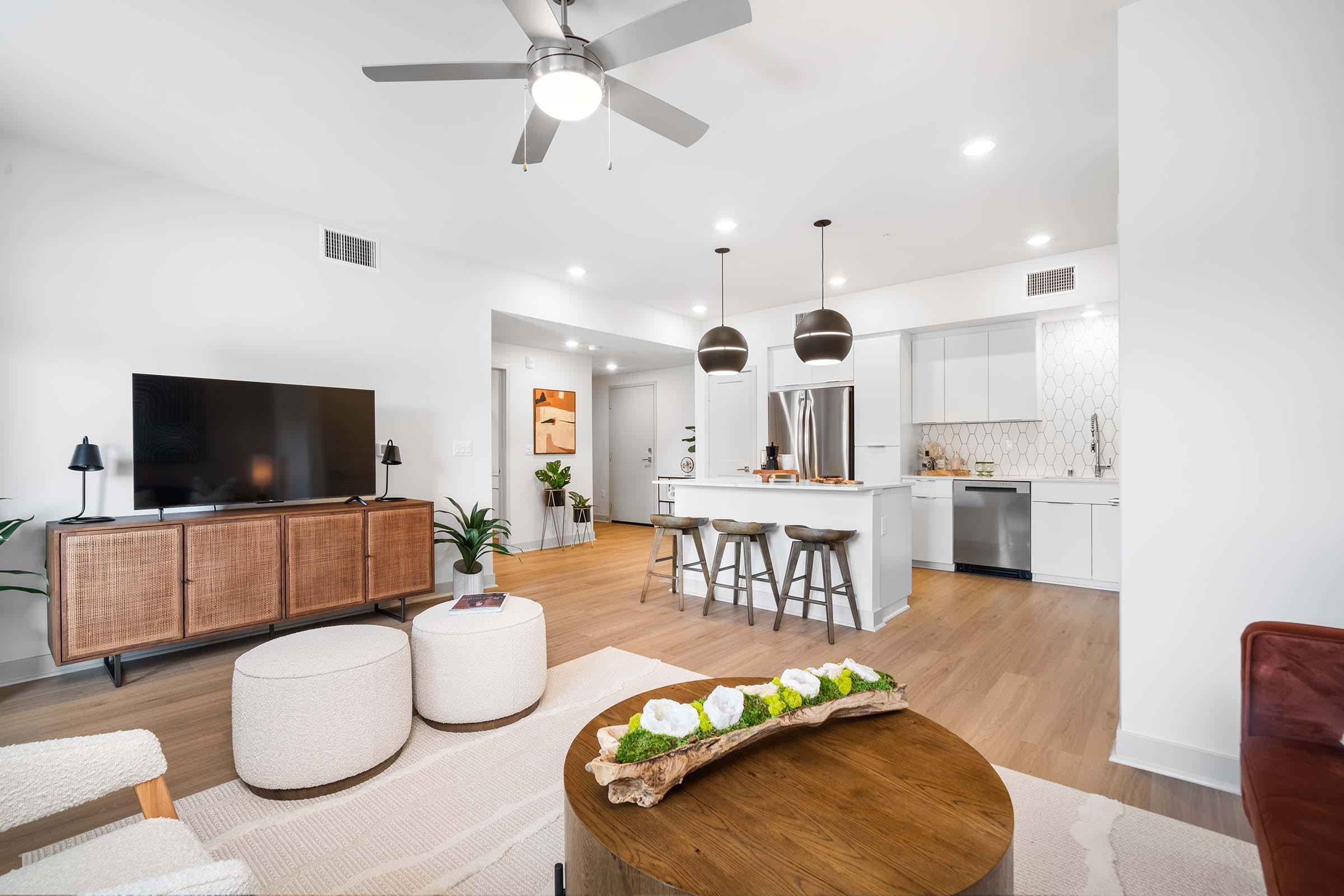 Modern living room featuring a television on a wooden console, two round ottomans, and a coffee table with a decorative centerpiece. A sleek kitchen with white cabinets and stainless steel appliances is visible in the background, alongside a dining area with minimalist decor and indoor plants.